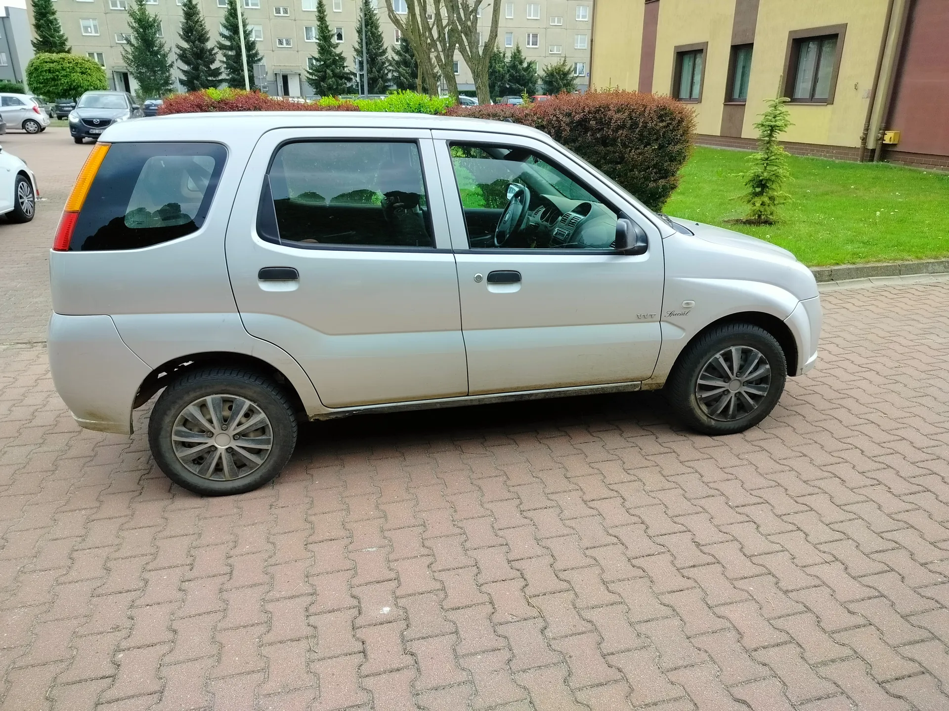 Srebrny samochód osobowy typu SUV zaparkowany na brukowanej powierzchni. Po lewej stronie widoczne są inne zaparkowane pojazdy, w tle budynki mieszkalne, drzewa iglaste i krzewy. Po prawej stronie trawnik oraz niewielkie drzewko. Samochód ma czarne felgi, ciemne szyby i widoczne ślady brudu na dolnej części nadwozia. W tle również zielone krzewy i drzewa liściaste.