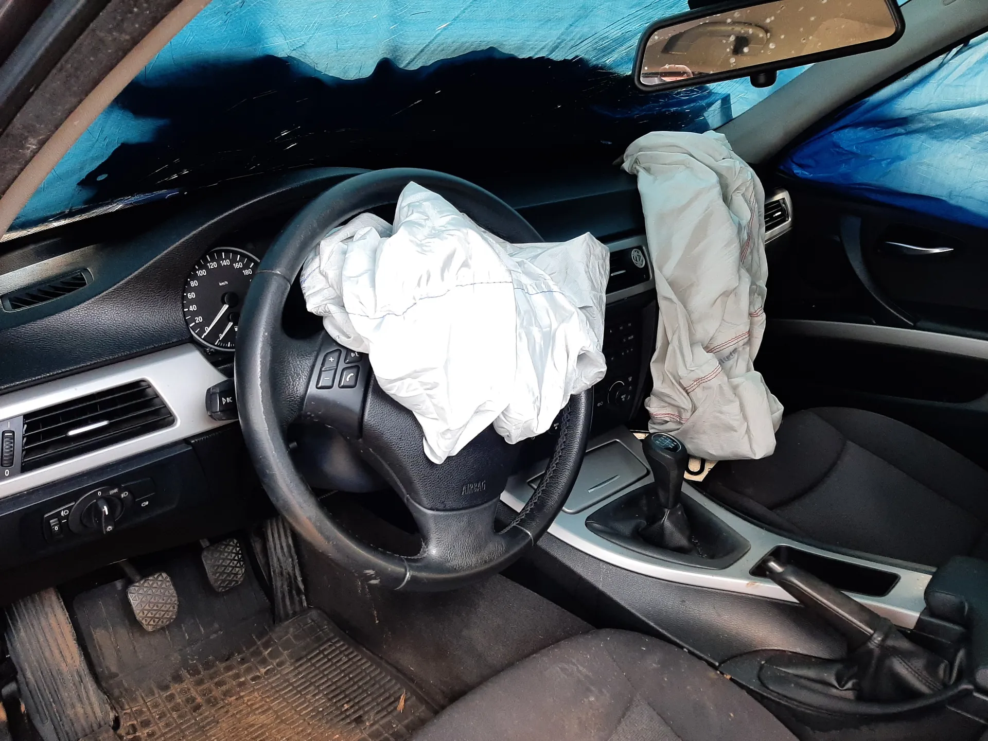 Wnętrze samochodu z rozwiniętymi poduszkami powietrznymi. Widoczna czarna kierownica z napisem AIRBAG, zegar prędkościomierza z zakresem do 180 km/h, dźwignia zmiany biegów, czarne siedzenia oraz brudne dywaniki podłogowe. Przez przednią szybę widoczny niebieski materiał. W lusterku wstecznym odbicie części otoczenia.