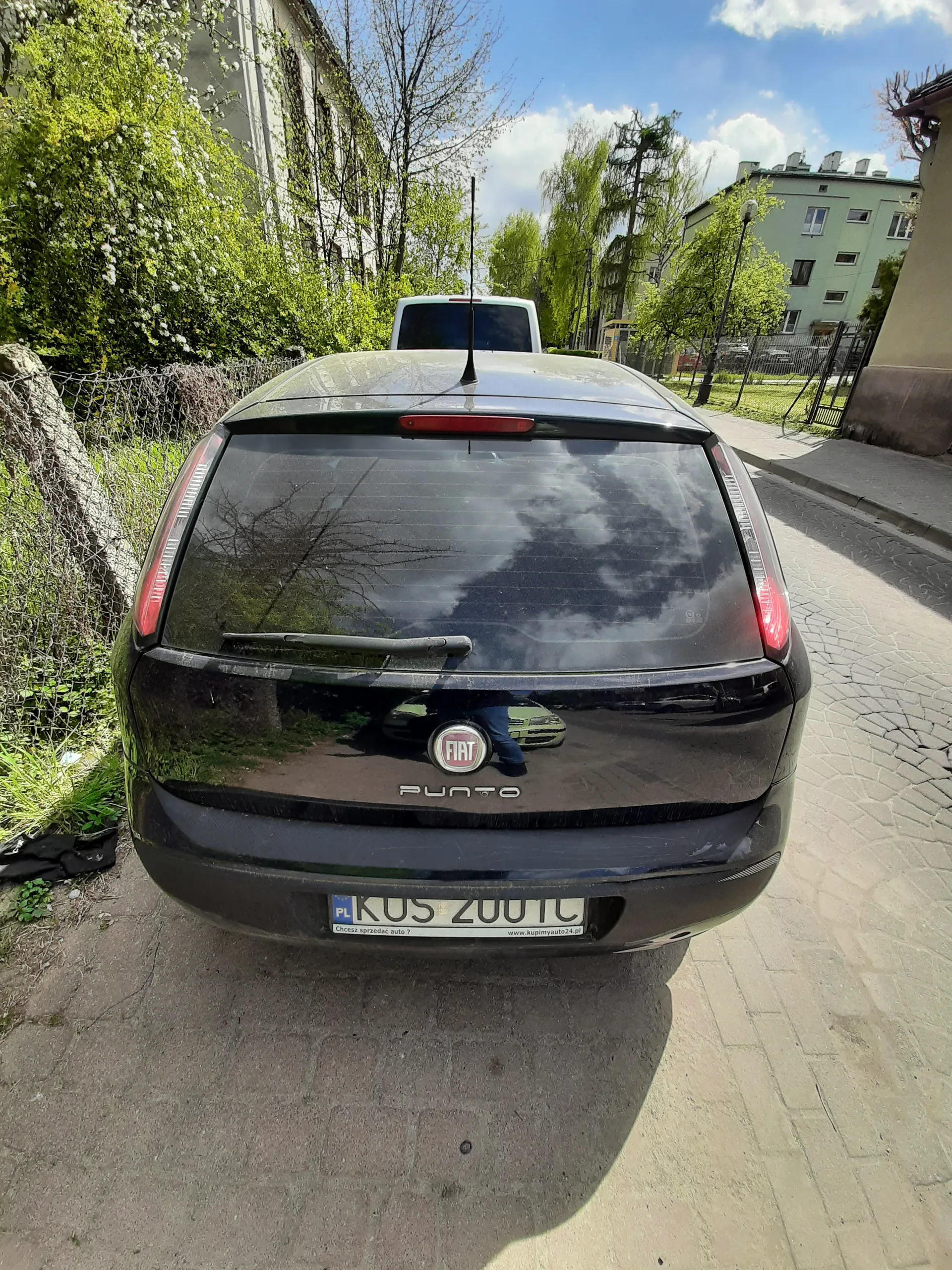Czarny samochód Fiat Punto z tyłu, zaparkowany na brukowanej ulicy. Widoczna tablica rejestracyjna z polskim kodem PL i numerem KUS 2007. W tle budynki mieszkalne, drzewa i niebo z chmurami. Na szybie tylnej odbicie chmur i drzew.