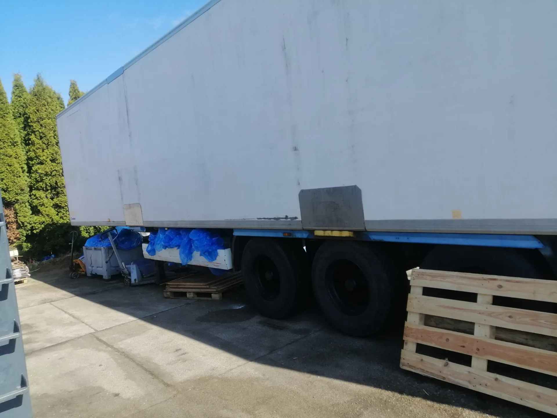 Boczny widok białej naczepy ciężarówki zaparkowanej na betonowym placu. Pod naczepą znajdują się niebieskie worki z materiałem oraz drewniane palety. W tle widoczne są zielone drzewa iglaste i bezchmurne, niebieskie niebo.