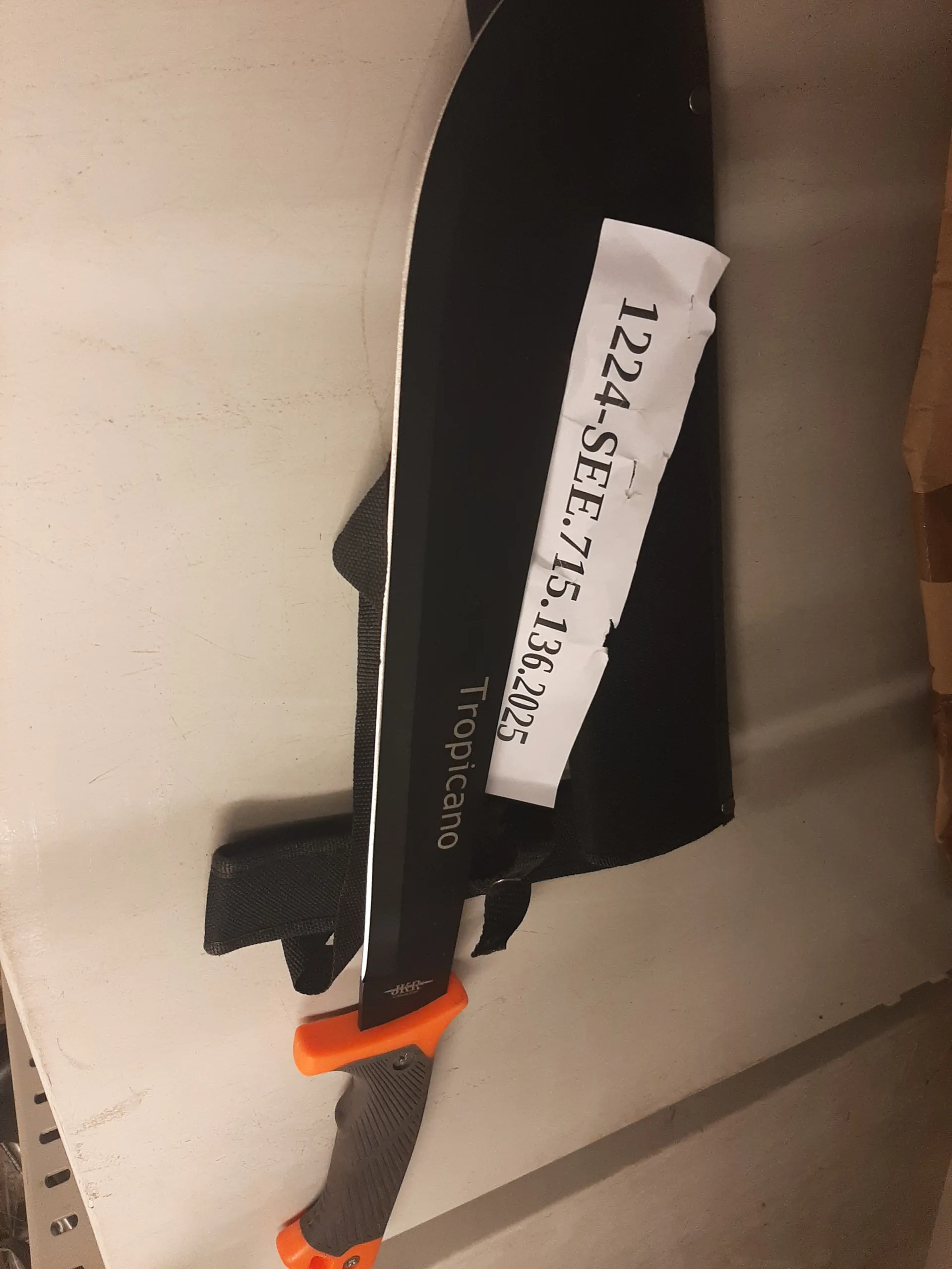 Czarny nóż typu machete z pomarańczowo-czarną rękojeścią leżący na czarnym pochwie. Na ostrzu widnieje napis 'Tropicano'. Na białej taśmie przyklejonej do ostrza znajduje się tekst '1224-SEE.715.136.2025'. Nóż umieszczony jest na jasnej powierzchni.