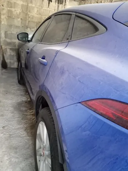 Niebieski samochód osobowy zaparkowany obok betonowej ściany z bloków. Widoczna tylna część pojazdu, czerwone światło tylnego reflektora, boczne lustro wsteczne oraz koła. Podłoga wykonana z betonu.