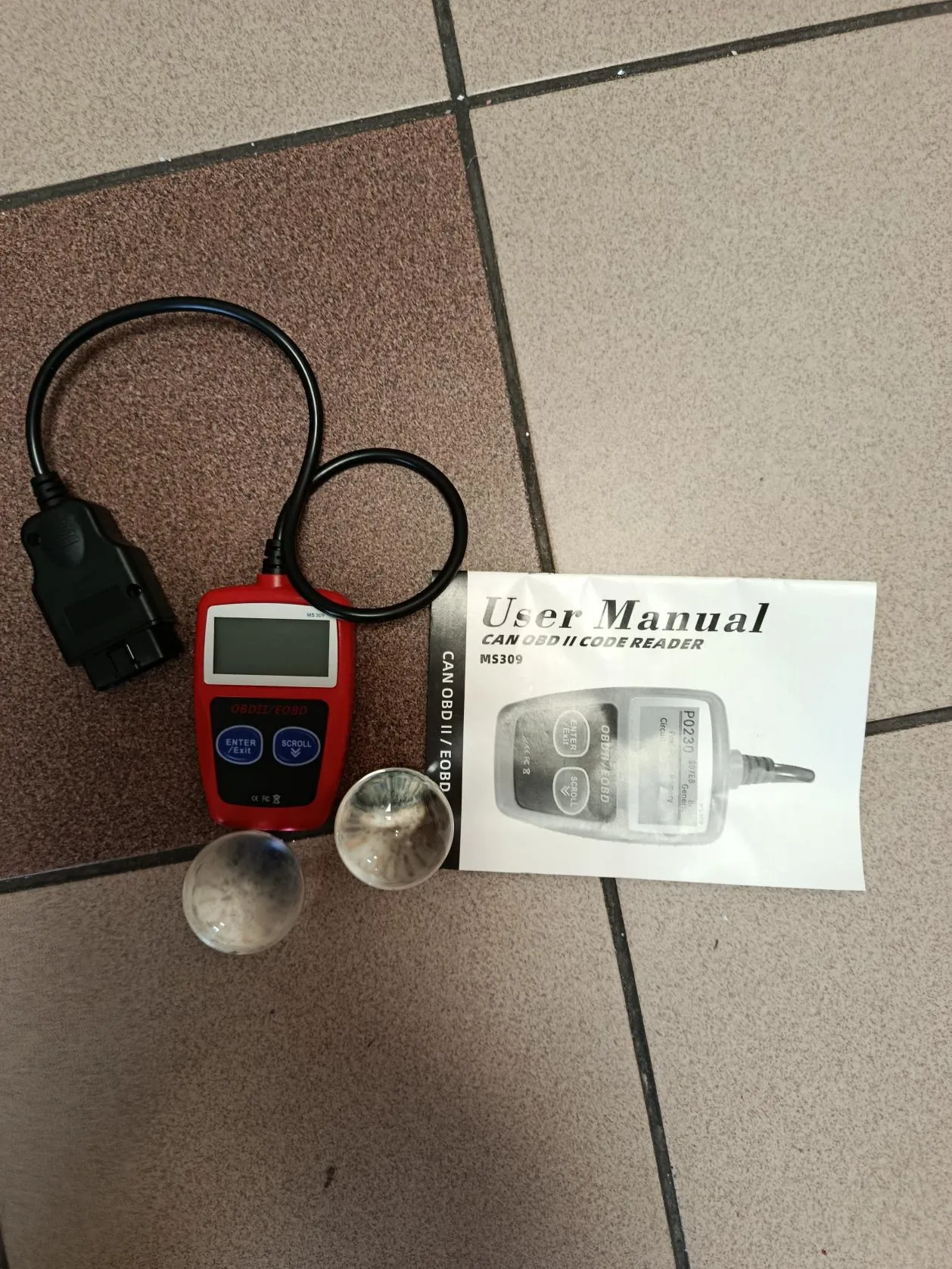 Czerwony czytnik kodów OBDII/EOBD z czarnym kablem i złączem, obok niego biała instrukcja użytkownika z napisem 'User Manual CAN OBD II CODE READER MS309' oraz dwie przezroczyste kulki. Wszystko leży na podłodze z kafelkami w kolorze brązowym i beżowym. Na urządzeniu widoczne przyciski 'ENTER/EXIT' i 'SCROLL' oraz wyświetlacz.