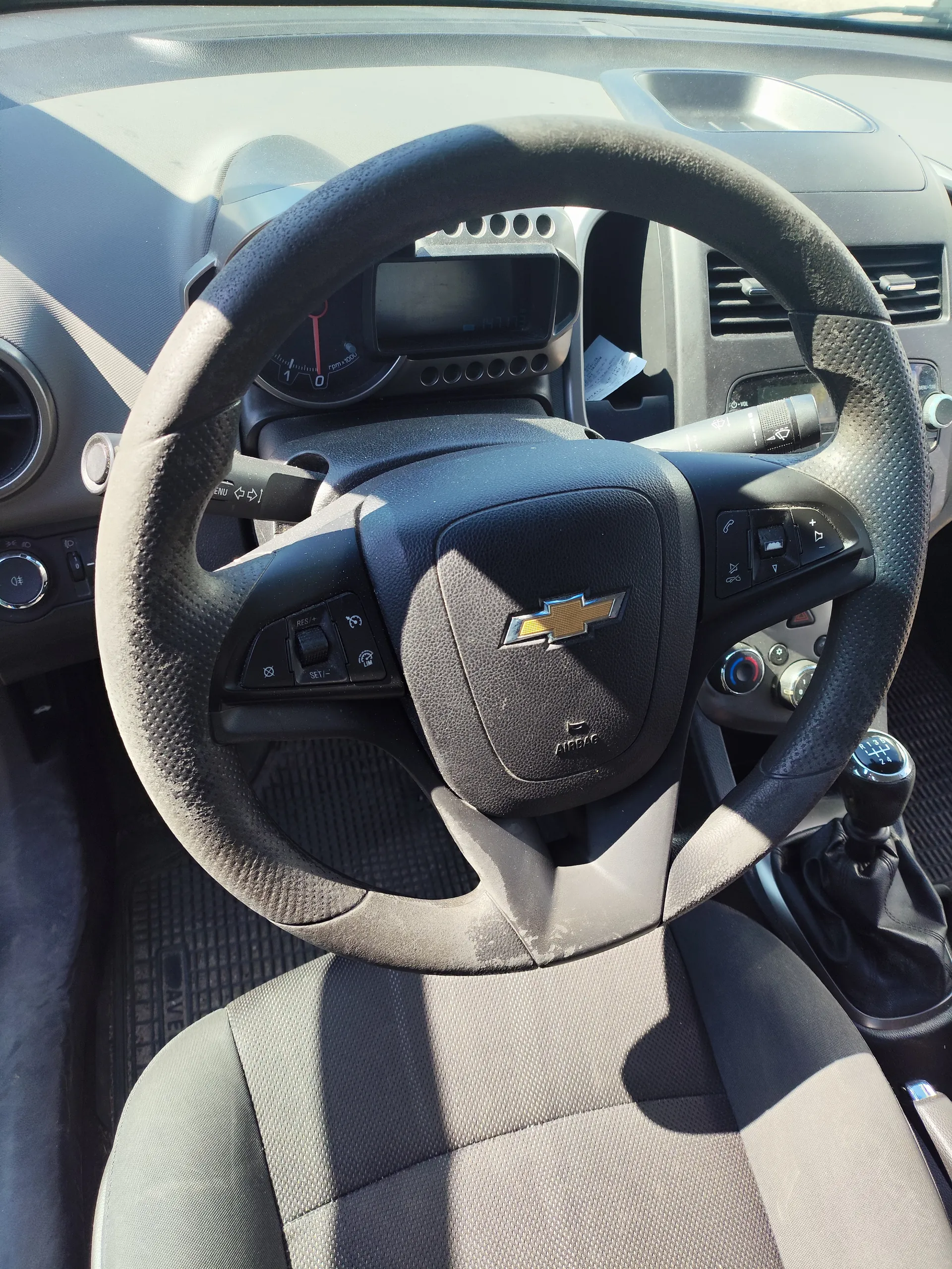 Wnętrze samochodu z widocznym kierownicą marki Chevrolet. Kierownica czarna, z przyciskami sterującymi po obu stronach i logo Chevrolet w centrum. Za kierownicą zegary samochodowe, w tym wskaźnik prędkości z czerwoną igłą. Po prawej stronie widoczna dźwignia zmiany biegów, a na desce rozdzielczej wentylatory i inne elementy sterujące. Siedzenia pokryte szarą tkaniną. Wnętrze w odcieniach czerni i szarości, oświetlone światłem słonecznym rzucającym cienie.