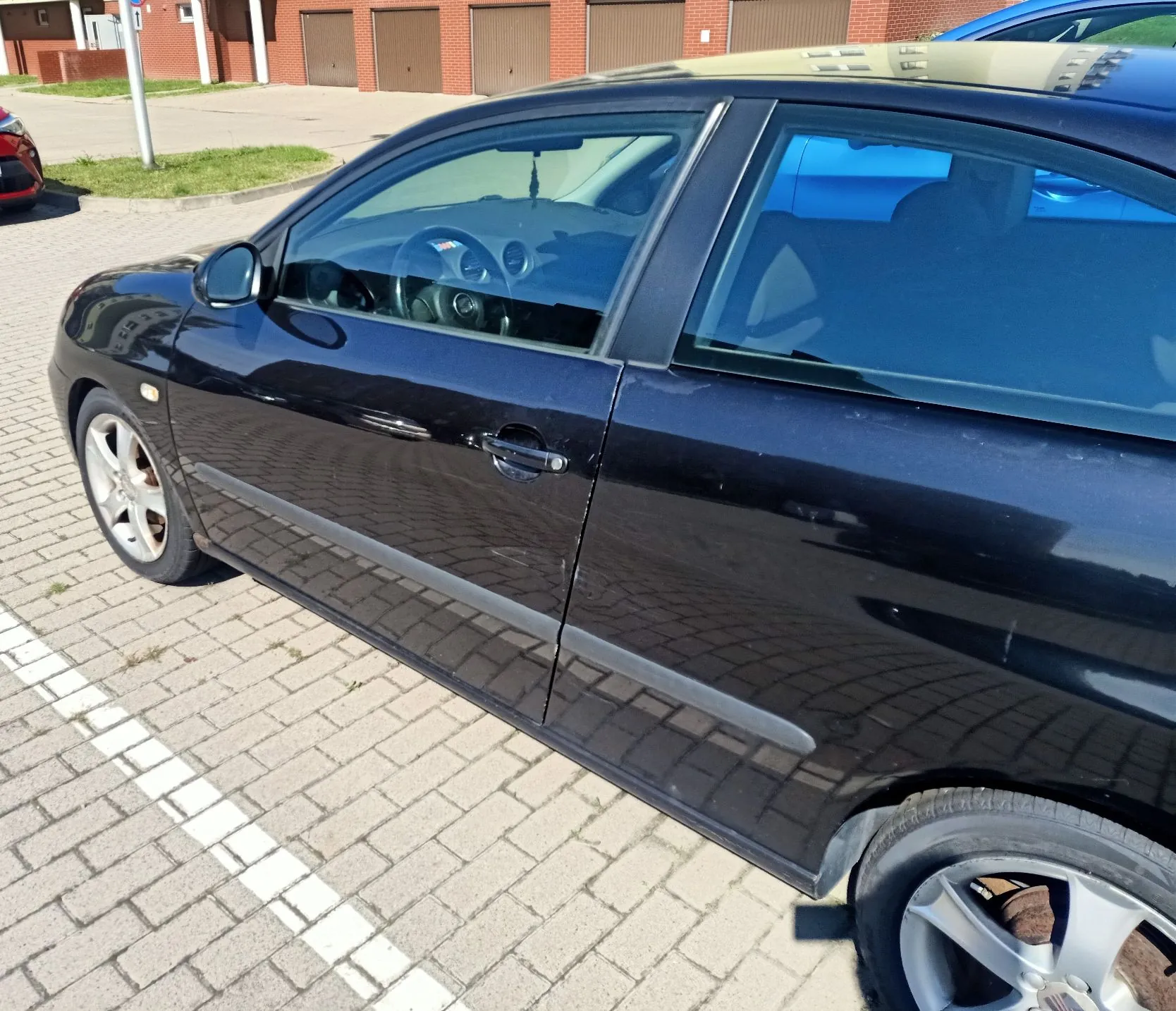 Zbliżenie na prawy bok czarnego samochodu osobowego zaparkowanego na kostce brukowej. Widoczne są przednie i tylne drzwi, lusterko boczne oraz dwa koła z aluminiowymi felgami. W tle widać fragmenty budynków z czerwonej cegły z brązowymi bramami garażowymi oraz fragment innego, czerwonego pojazdu po lewej stronie.