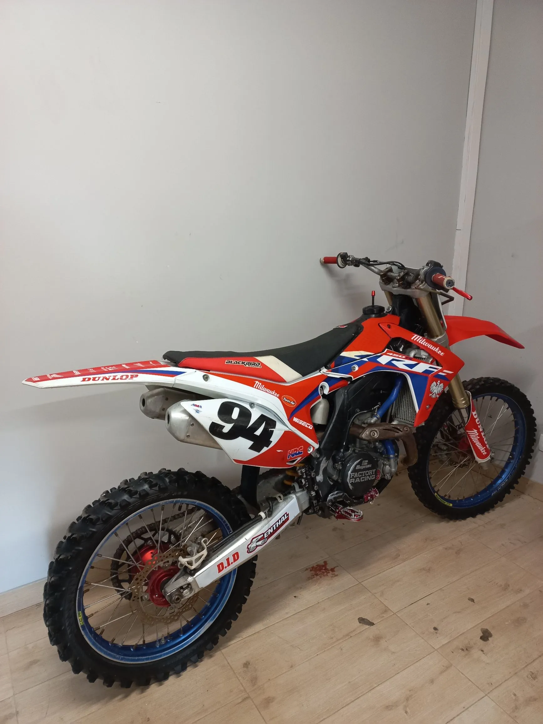 Motocykl crossowy w kolorach czerwonym, białym i niebieskim, zaparkowany w pomieszczeniu na jasnej drewnianej podłodze na tle białej ściany. Motocykl posiada numer 94 na bocznej owiewce oraz liczne napisy sponsorskie, m.in. 'Milwaukee', 'Dunlop', 'D.I.D' i 'Renthal'. Posiada terenowe opony z wyraźnym bieżnikiem.