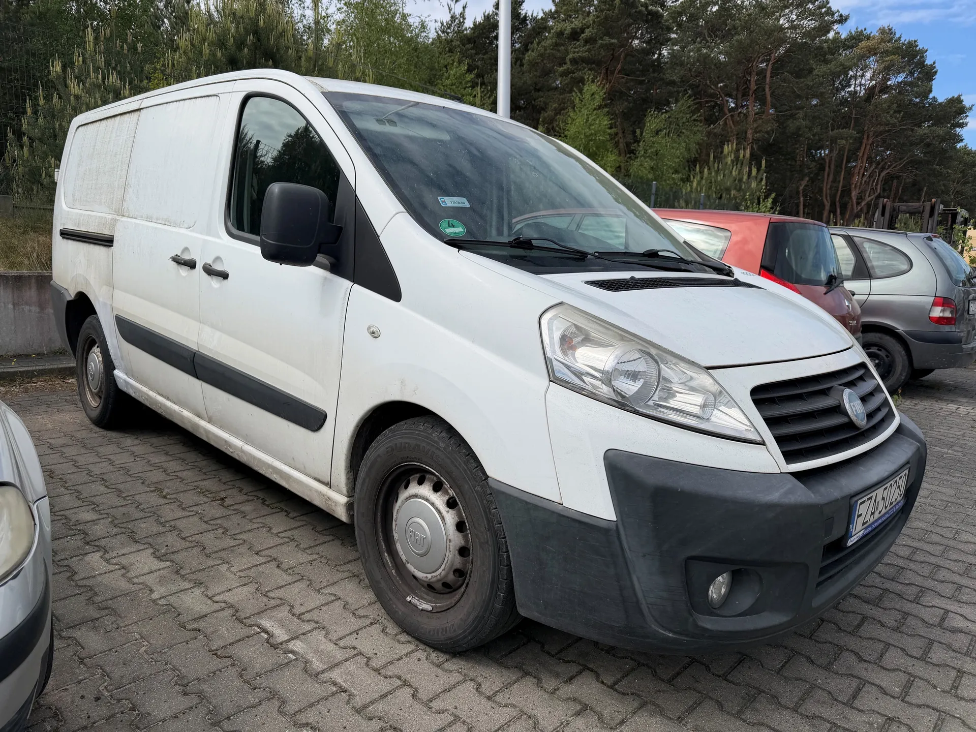 Biały furgon marki Fiat z czarną listwą boczną i tablicą rejestracyjną EZA 50250, zaparkowany na brukowanym placu. W tle widoczne są inne pojazdy: czerwony i szary samochód, a także drzewa i ogrodzenie. Na przedniej szybie furgona znajdują się dwa naklejki.