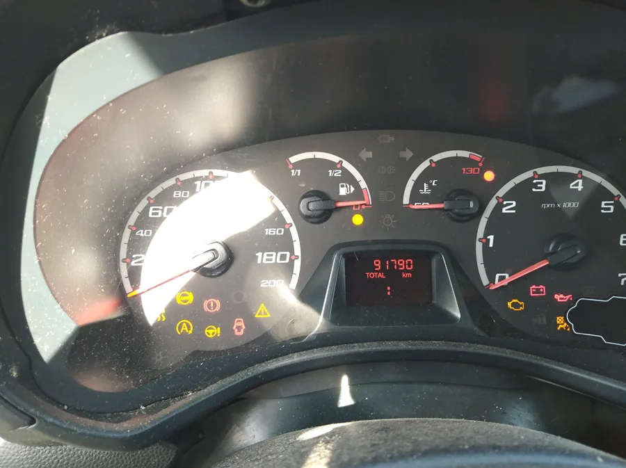 Tablica rozdzielcza samochodu z licznikami: prędkościomierz (0-200 km/h), wskaźnikiem paliwa (połowa zbiornika), termometrem chłodniczym, tachometrem (0-7000 obr./min) oraz wyświetlaczem cyfrowym pokazującym 91790 km. Widoczne są żółte i czerwone lampki ostrzegawcze, w tym wskaźnik paliwa, bateria, olej i inne. Część kierownicy widoczna u dołu. Wyświetlacz i znaki są czytelne, bez oznak nieprawidłowej orientacji.