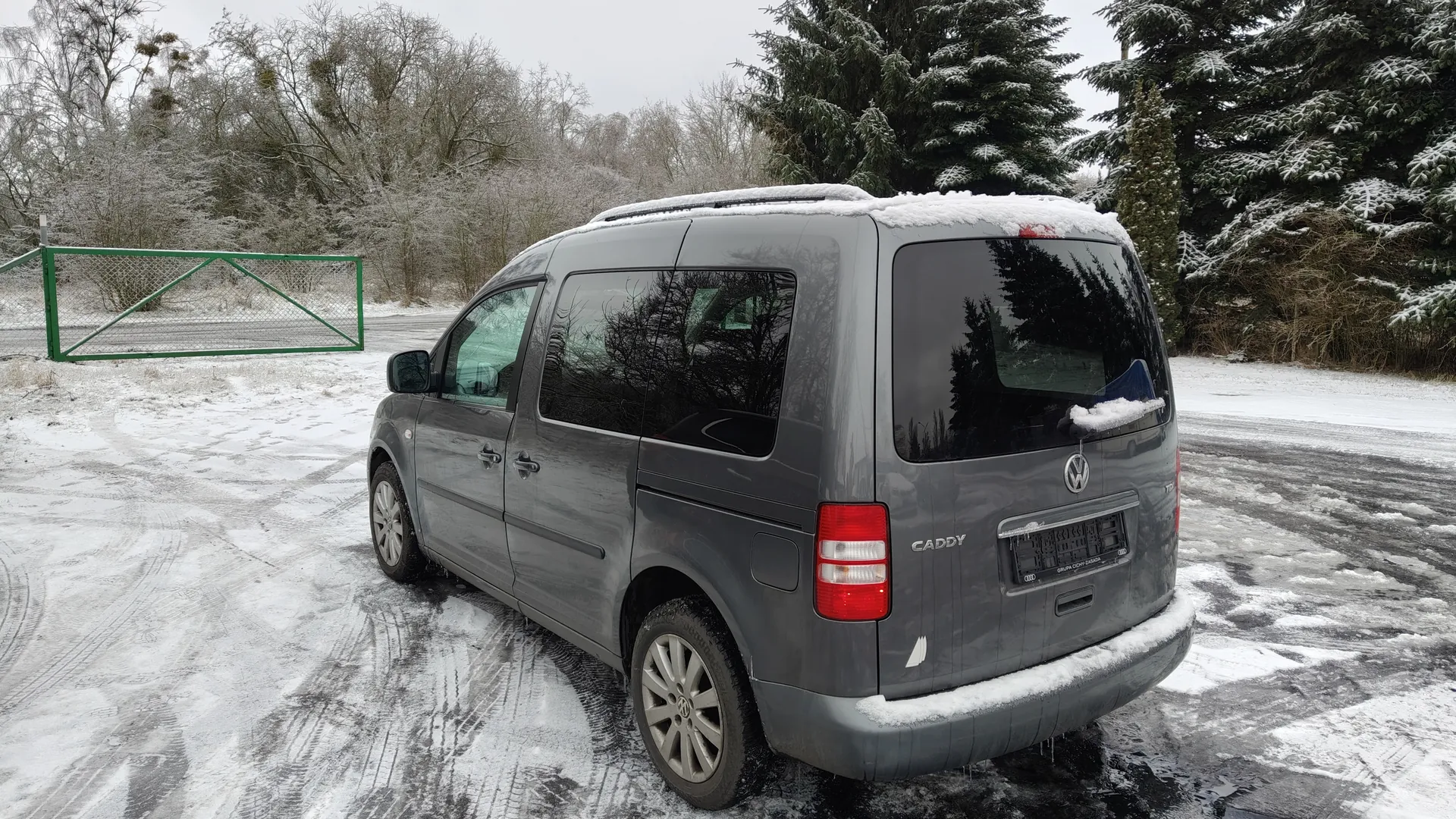 Szary samochód dostawczy Volkswagen Caddy zaparkowany na śnieżnej drodze. Na dachu i tylnym zderzaku widoczny śnieg. W tle zielona metalowa brama, drzewa pokryte śniegiem oraz odciski opon na drodze. Na tylnym zderzaku widoczny napis "CADDY" oraz logo Volkswagena.
