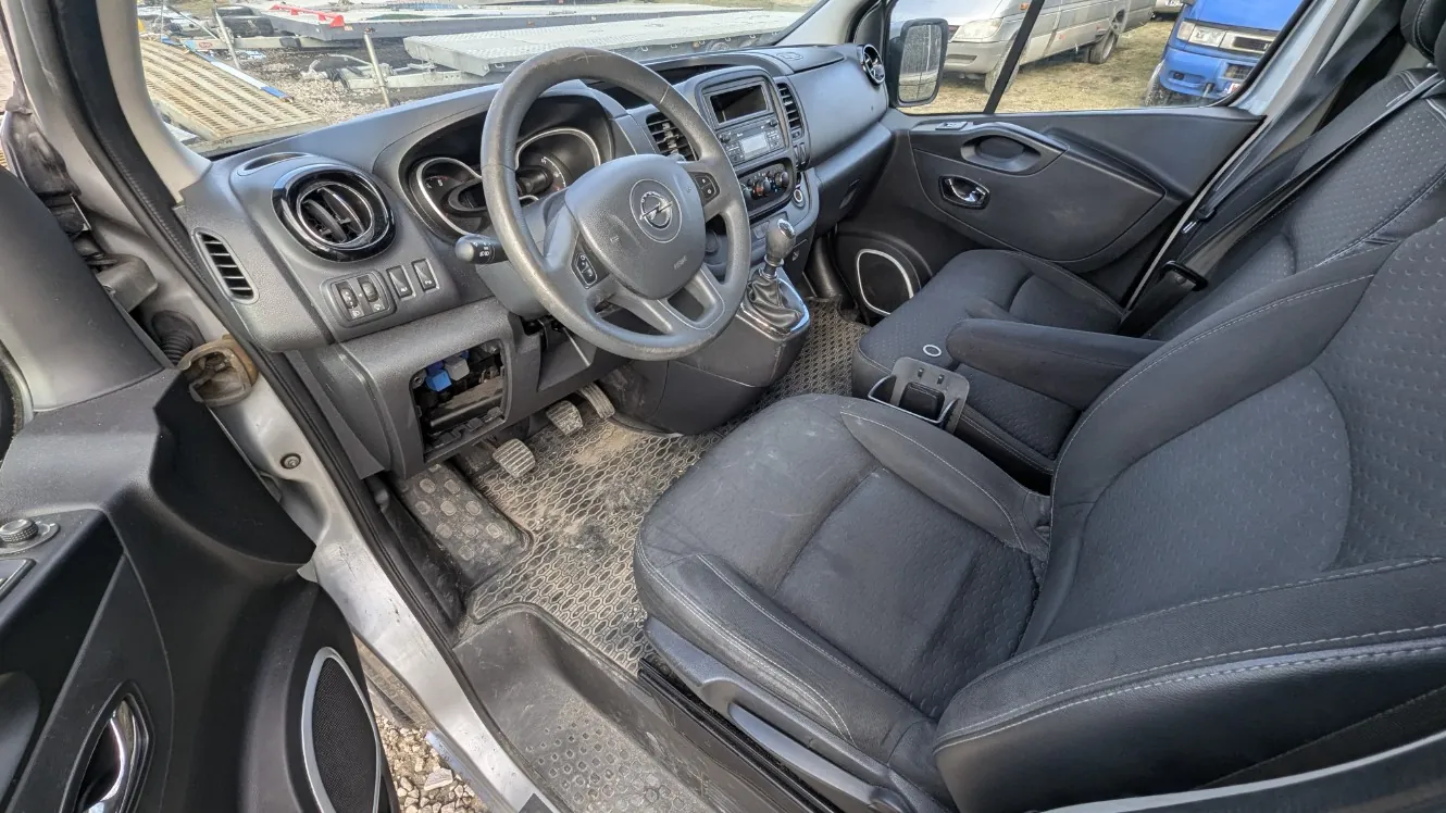 Wnętrze pojazdu widziane z otwartego drzwi pasażera. Widoczna kierownica z logo, deska rozdzielcza z wentylacjami, przyciskami i ekranem, czarne siedzenia, dźwignia zmiany biegów oraz brudne dywaniki. Na zewnątrz przez otwarte drzwi widać inne pojazdy i teren z materiałem budowlanym.