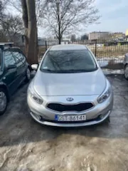 Srebrny samochód marki Kia zaparkowany na zamarzniętym podłożu. Widoczna tablica rejestracyjna z białym tekstem 'S5141' na niebieskim tle. Po lewej stronie częściowo widoczny ciemny samochód. W tle drzewa bez liści, metalowe ogrodzenie oraz budynki. Niebo jest jasne.