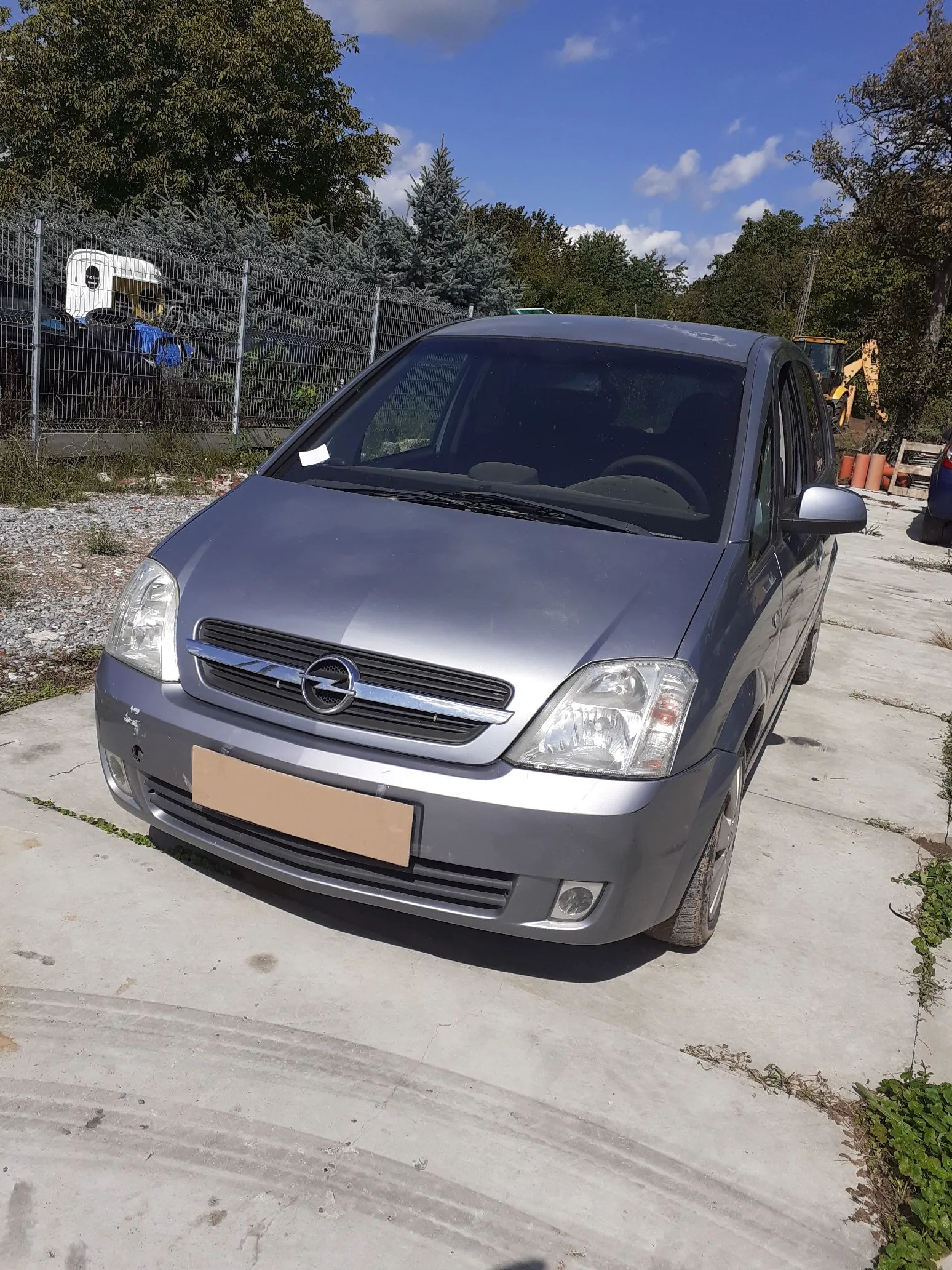 Srebrny samochód marki Opel zaparkowany na betonowym podjeździe. Pojazd jest widoczny z przodu pod kątem. W tle znajduje się metalowy płot, zielone drzewa oraz fragment żółtej maszyny budowlanej pod niebem z lekkimi chmurami.