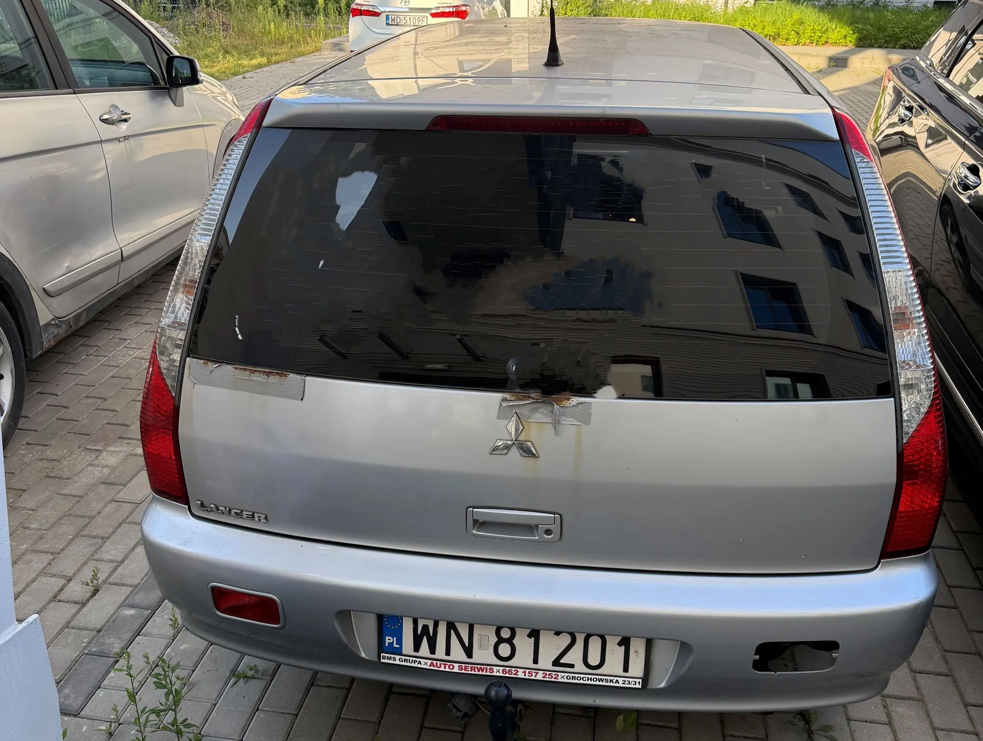Srebrny samochód marki Mitsubishi Lancer widziany od tyłu. Pojazd posiada tablicę rejestracyjną o numerze WN 81201. Na ramce tablicy widnieje napis: 'BMS GRUPA » AUTO SERWIS » 662 157 252 » GROCHOWSKA 23/31'. Na klapie bagażnika znajduje się logo Mitsubishi oraz napis LANCER po lewej stronie. Karoseria w okolicach logo jest lekko uszkodzona i zmatowiała.