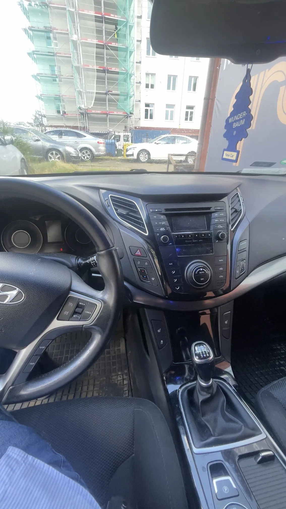 Widok z wnętrza samochodu marki Hyundai, z perspektywy kierowcy. Widoczna jest kierownica z logo producenta, deska rozdzielcza z radiem i panelami sterowania klimatyzacją oraz lewarek zmiany biegów manualnej. Przez przednią szybę widać parking z zaparkowanymi samochodami oraz w oddali budynek w trakcie budowy, osłonięty zieloną siatką budowlaną. Na lusterku wstecznym wisi odświeżacz powietrza Wunder-Baum w kształcie choinki.