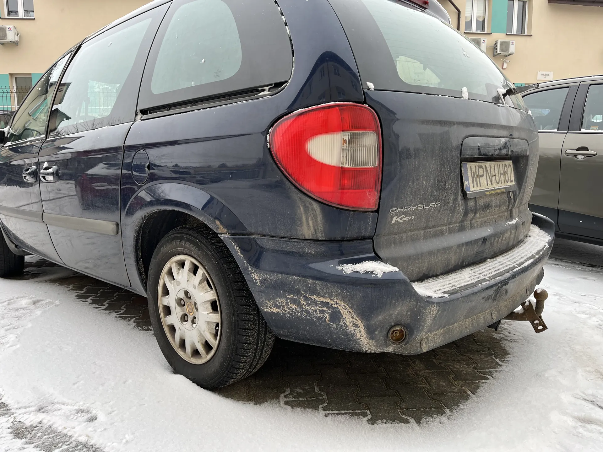 Niebieski samochód osobowy Chrysler Voyager z tablicą rejestracyjną WPN UH82 zaparkowany na śnieżnej nawierzchni. Na karoserii, zderzaku i dachu ślady śniegu i brudu, czerwone światła tylne, srebrne felgi. W tle budynek z klimatyzatorami i szary pojazd.