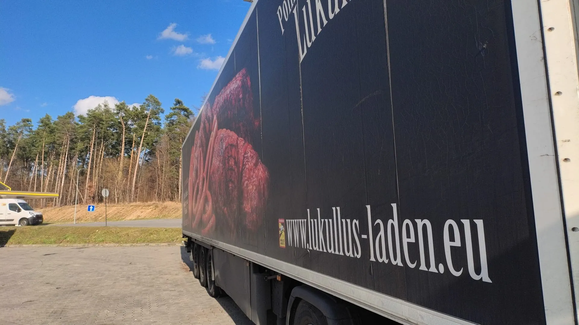 Czarna naczepa ciężarówki z dużym obrazem mięsa i widocznym tekstem 'www.lukullus-laden.eu' oraz częściowo widocznym napisem 'Pork Lukullus'. W tle niebo z chmurami, las iglasty, droga, biały samochód dostawczy oraz znaki drogowe. Całość na tle jasnego dnia.