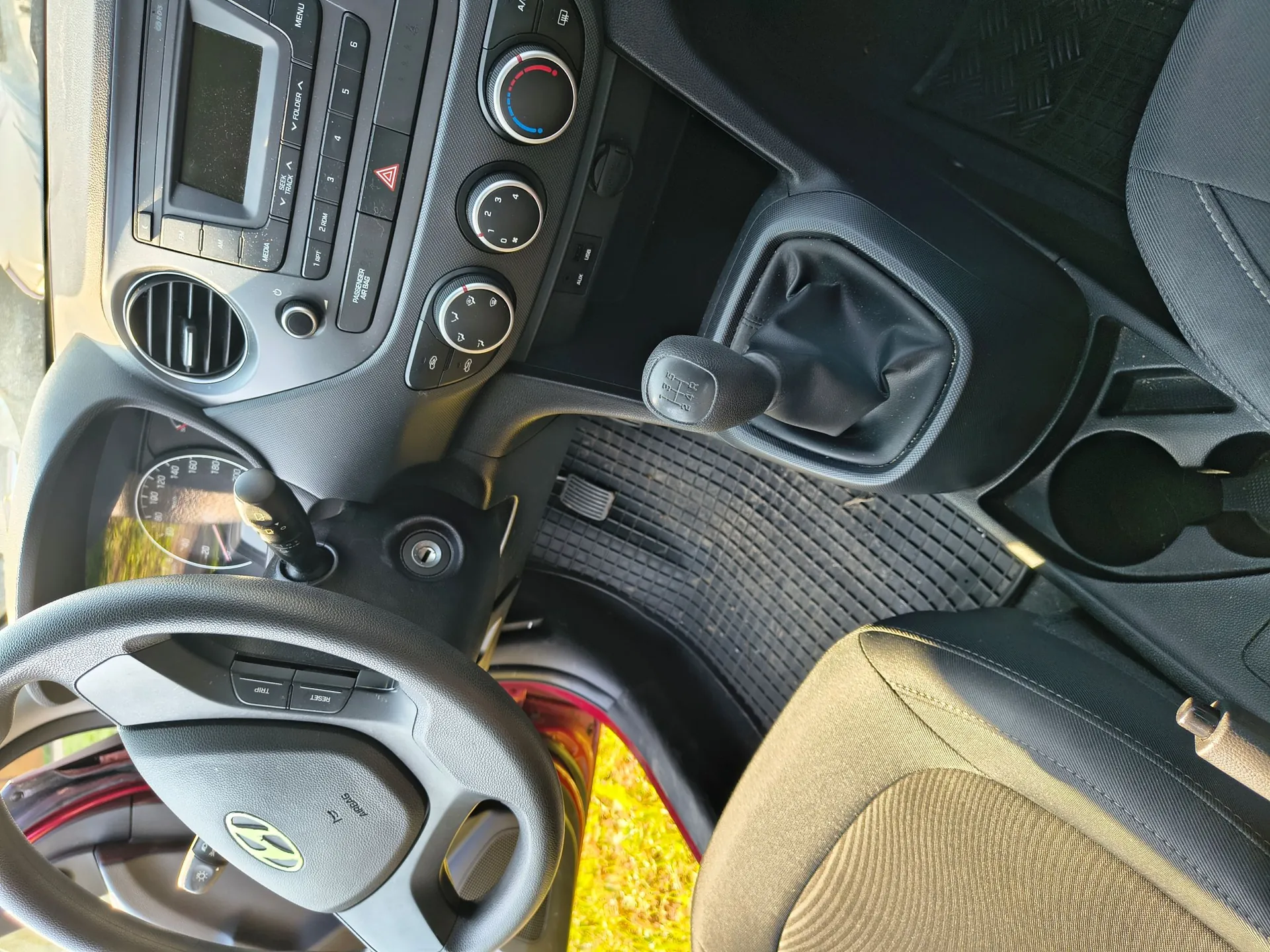 Widok z góry na wnętrze samochodu marki Hyundai. Widoczna jest kierownica z logo producenta, deska rozdzielcza z radiem, przyciskami sterowania i nawiewami, dźwignia zmiany biegów w czarnej skórze oraz przedni fotel pasażera. Na podłodze znajduje się gumowa mata z wypustkami.