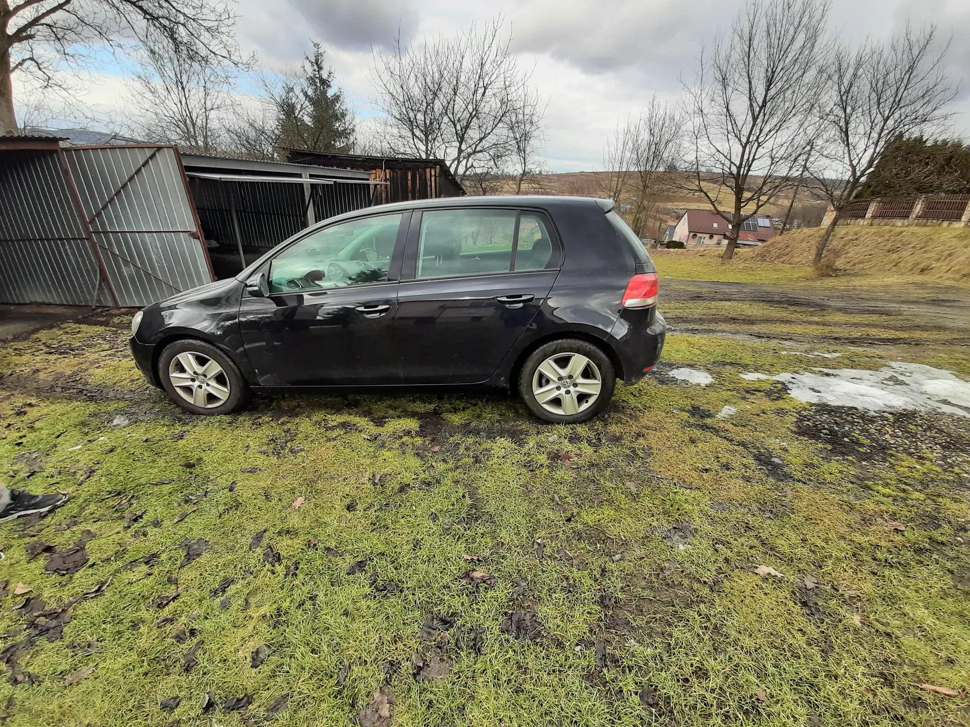 Czarny samochód osobowy typu hatchback zaparkowany na trawiastym obszarze z resztkami śniegu. W tle metalowe garaże, bezlistne drzewa, domy i chmurne niebo. Na ziemi rozrzucone są suche liście.