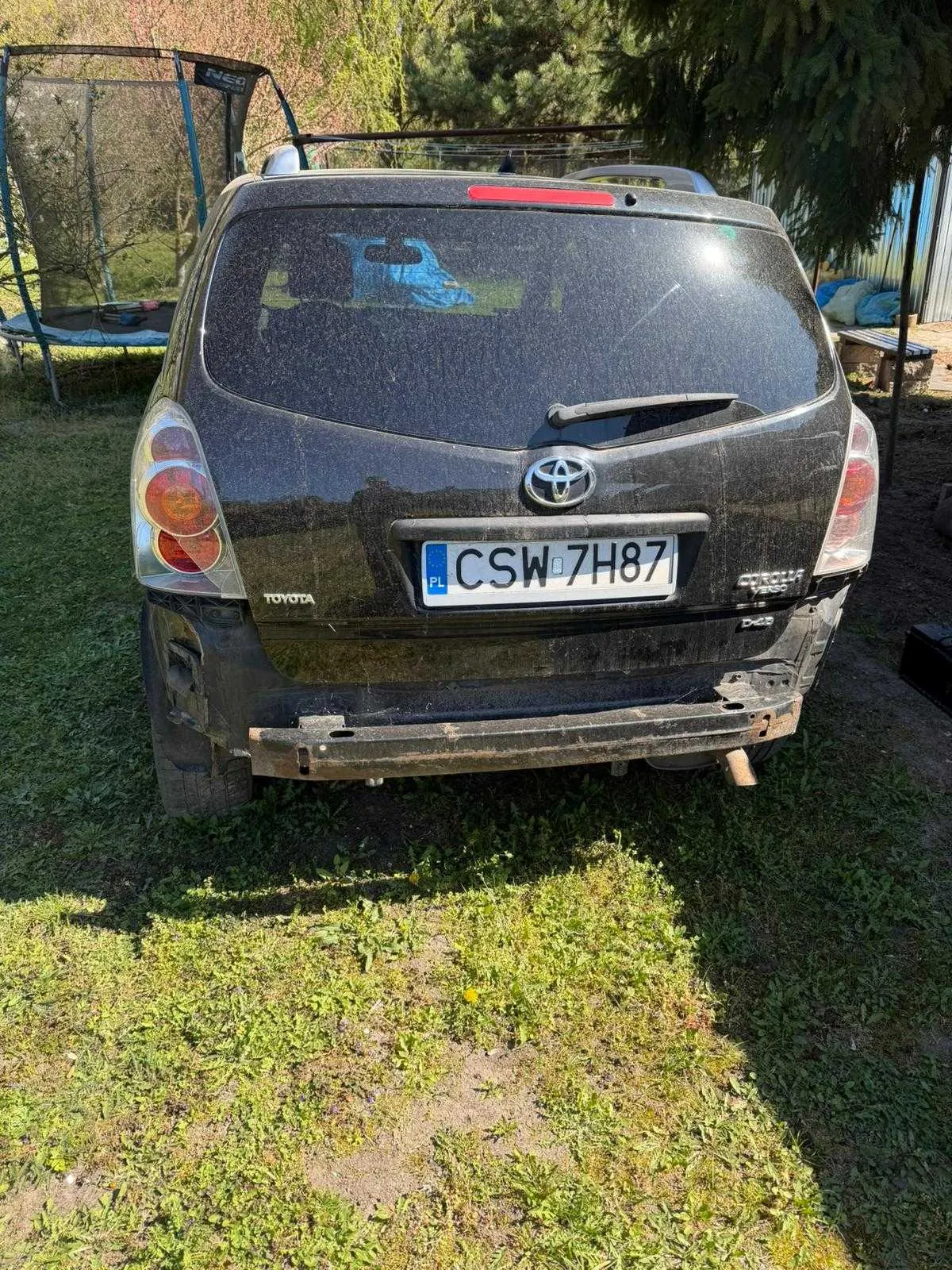 Z tyłu czarny samochód marki Toyota z tablicą rejestracyjną CSW 7H87 (PL). Samochód jest zabrudzony, z uszkodzonym tylnym zderzakiem. W tle trampolina, drzewa i budynek. Na trawie widoczne są zielone rośliny.