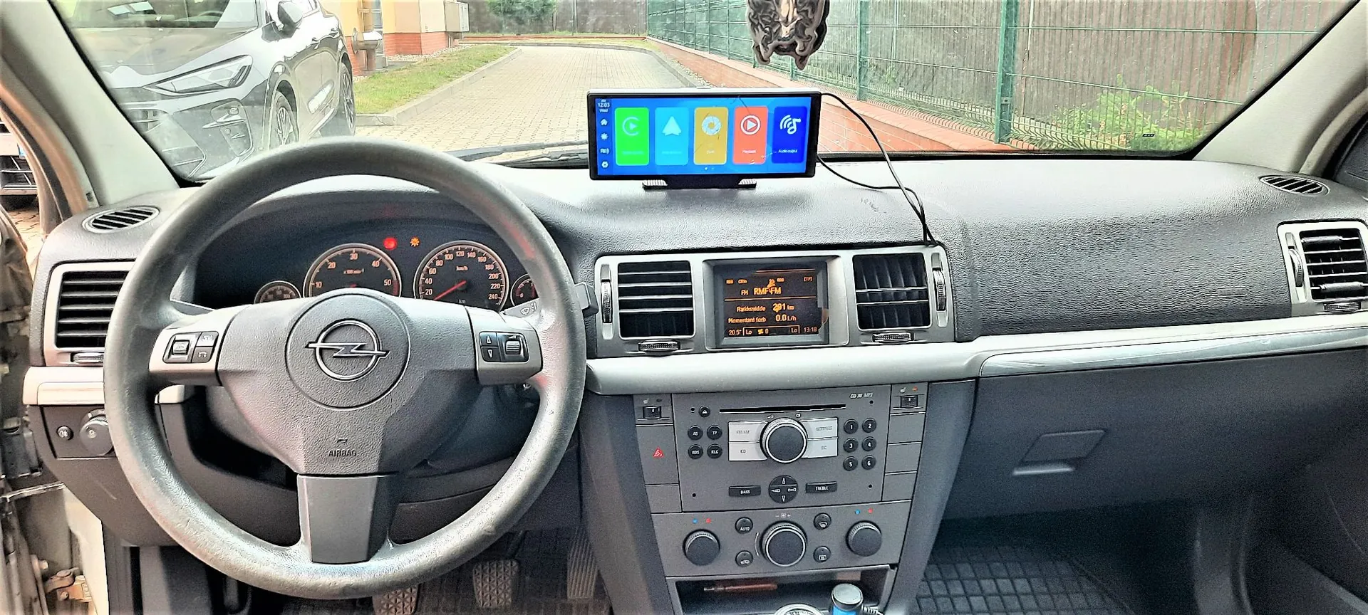 Wnętrze samochodu marki Opel widziane z perspektywy kierowcy. Widoczna jest kierownica z logo producenta, deska rozdzielcza z zegarami, system audio oraz klimatyzacja. Na górnej części deski rozdzielczej zamontowany jest dodatkowy ekran multimedialny wyświetlający kolorowe ikony aplikacji. Przez przednią szybę widoczny jest fragment innego srebrnego samochodu oraz podjazd z zielenią w tle.