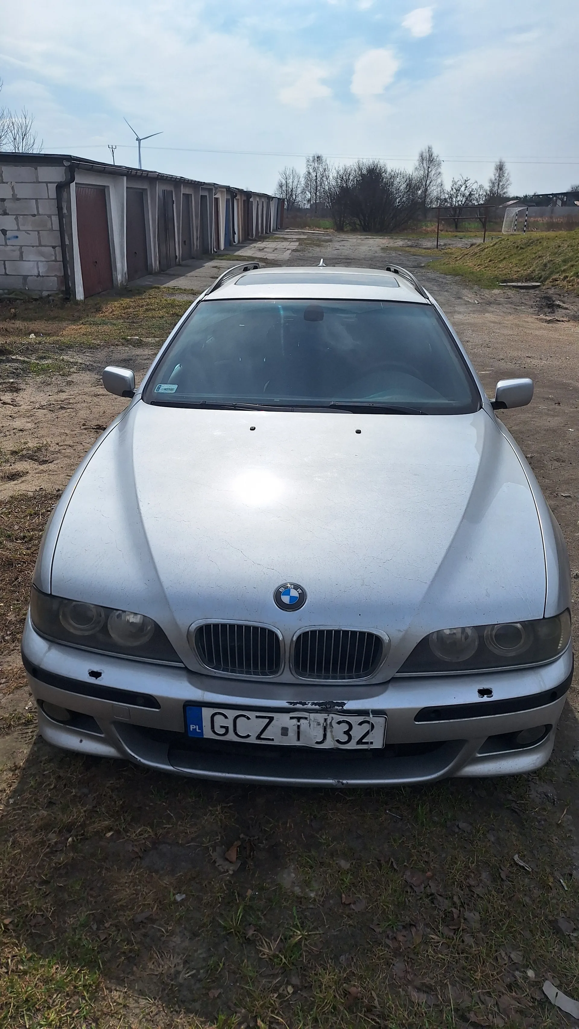 Srebrny samochód marki BMW z tablicą rejestracyjną GCZ TJ32 (PL) zaparkowany na brudnej drodze. W tle budynki garażowe, trawa, drzewa i niebo z chmurami. Samochód ma uszkodzenia na przednim zderzaku i widoczne jest logo BMW na masce.