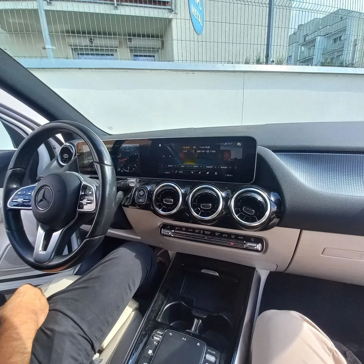 Widok z wnętrza samochodu na deskę rozdzielczą, kierownicę oraz przedni panel sterowania. Na kierownicy widoczne jest logo marki Mercedes-Benz. W centrum znajduje się szeroki ekran multimedialny wyświetlający menu radia i ustawienia pojazdu. Poniżej ekranu umieszczono trzy okrągłe nawiewy powietrza w chromowanych obramowaniach oraz panel z przyciskami sterowania klimatyzacją. W dolnej części zdjęcia widoczne są nogi osoby siedzącej na fotelu kierowcy w czarnych spodniach.
