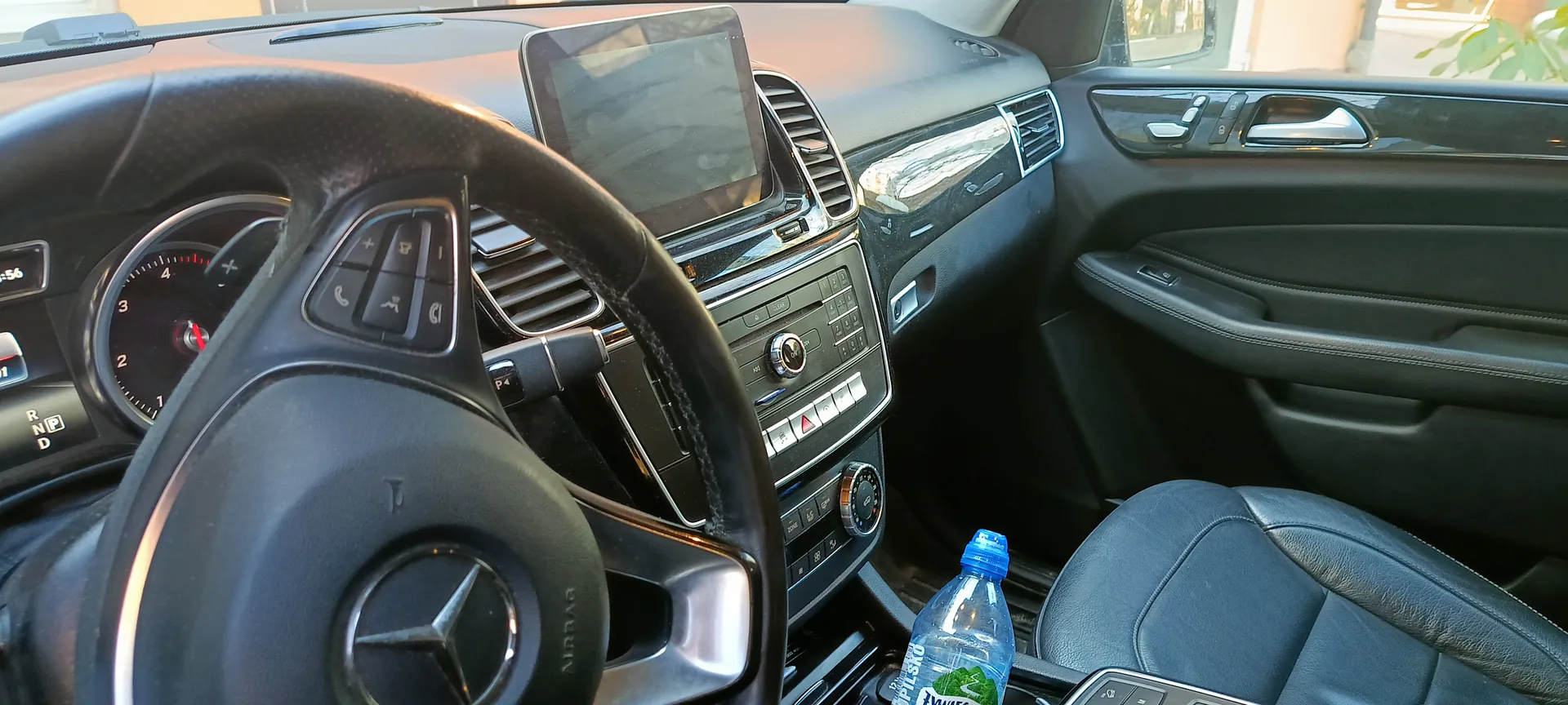 Wnętrze samochodu marki Mercedes z czarną kierownicą z logo trójgwiazdy, tablicą rozdzielczą z prędkościomierzem i wskaźnikiem biegów (R, N, P, D), centralnym ekranem multimedialnym, przyciskami sterującymi klimatyzacją i radiem. Na siedzeniu pasażera znajduje się plastikowa butelka z niebieskim korkiem i częściowo widocznym napisem PILSKO. Wnętrze wykończone czarną skórą, widoczne drzwi z uchwytem i przyciskami sterującymi szybami.