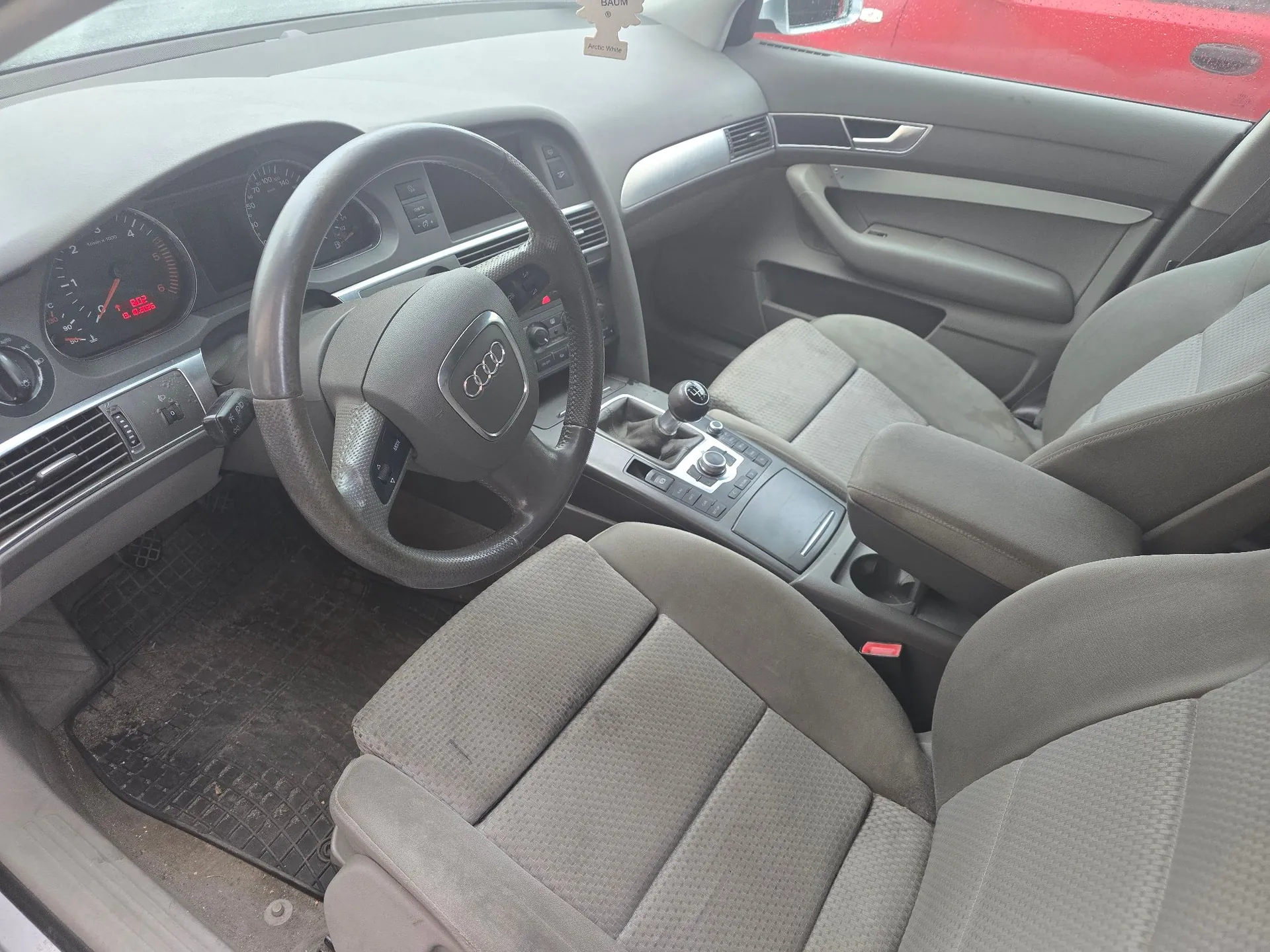 Wnętrze samochodu marki Audi widziane z perspektywy pasażera. Widoczna jest kierownica z logo Audi, deska rozdzielcza z zegarami, lewarek zmiany biegów, konsola środkowa oraz dwa przednie fotele tapicerowane szarym materiałem. Na podłodze znajduje się gumowa mata.
