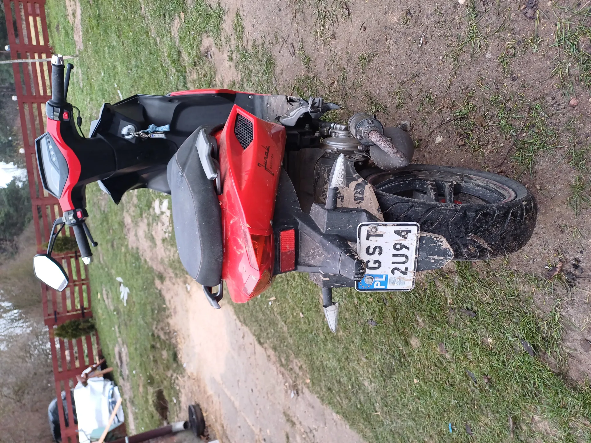 Czerwono-czarny skuter Juno z tablicą rejestracyjną PL GST 2U94, widok od tyłu. Brudne koło tylne, czarne siedzenie. Stojący na trawie i błotnej ścieżce. Tło: czerwone ogrodzenie, drzewa, rozrzucone przedmioty. Lusterka i elementy sterowania widoczne.