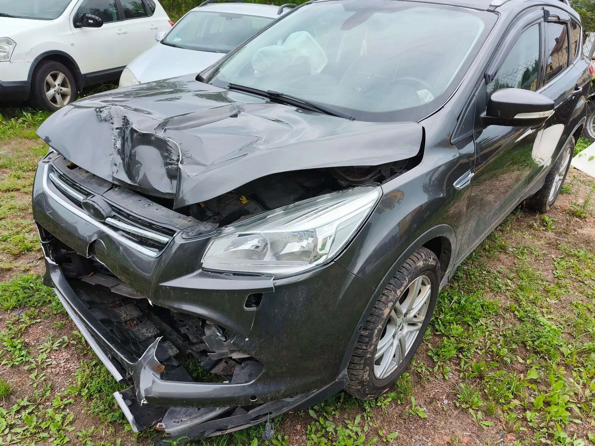 Zdjęcie przodu ciemnoszarego samochodu osobowego po kolizji. Maską jest mocno wgnieciona i wygięta do góry, przedni zderzak jest rozbity i częściowo oderwany od karoserii, a grill jest uszkodzony. Wewnątrz pojazdu, za przednią szybą, widoczna jest rozwinięta biała poduszka powietrzna kierowcy. Samochód stoi na trawiastym podłożu, w tle widoczne są dwa inne białe samochody.