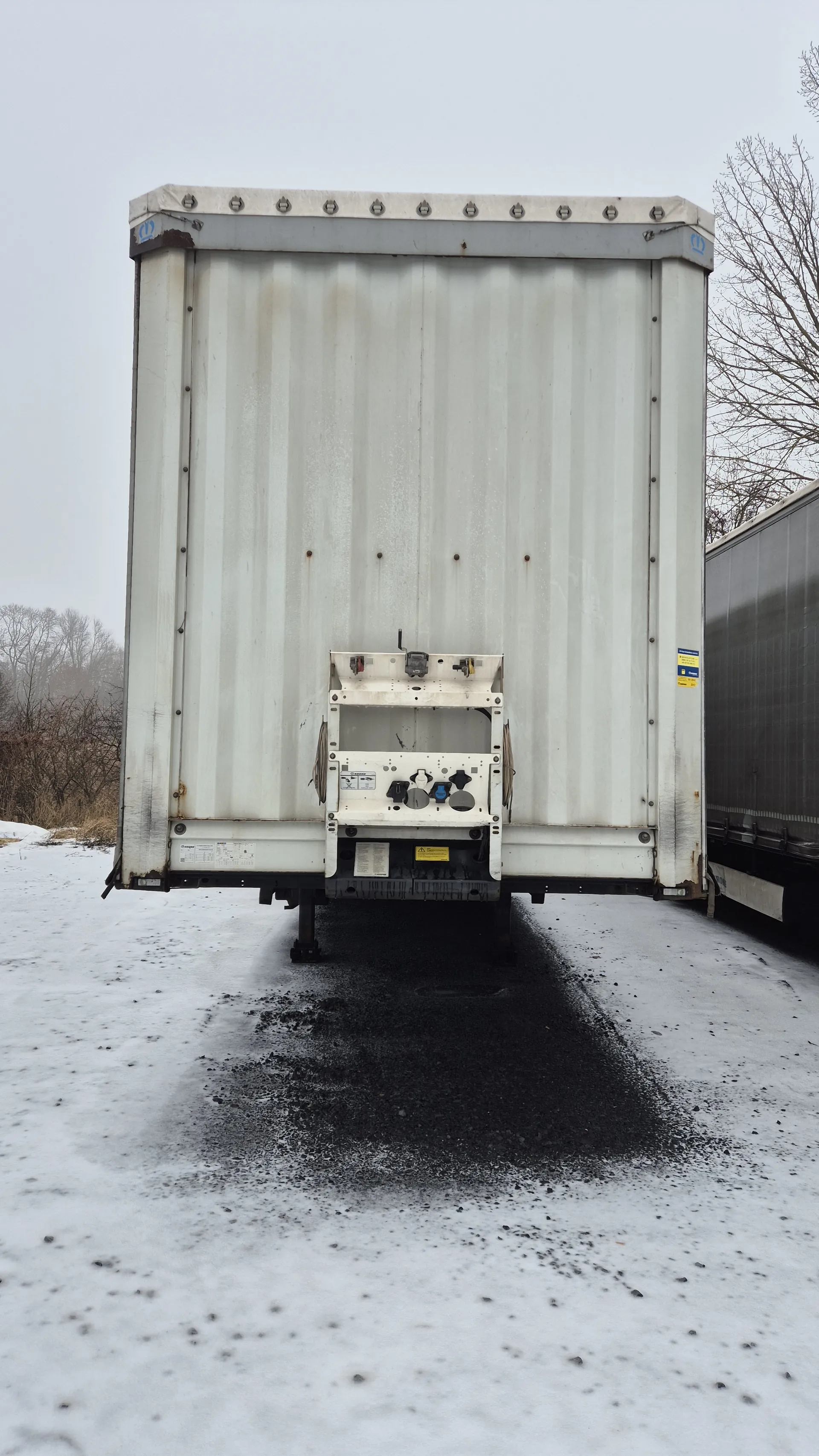 Biały chłodniczy kontener przyczepowy z widocznymi elementami sterującymi i etykietami. Kontener ma ślady rdzy oraz śruby po bokach. W tle: śnieżna powierzchnia, nagie drzewa i pochmurne niebo. Widoczna również część budynku.