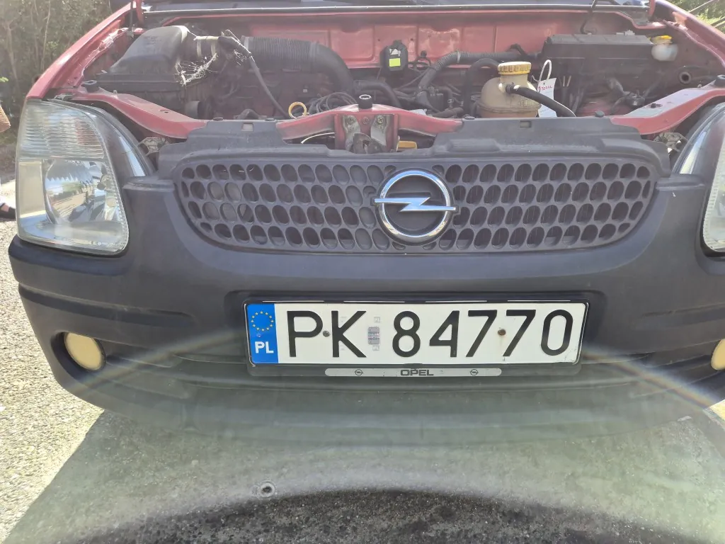 Zbliżenie na przód czerwonego samochodu marki Opel z otwartą maską, odsłaniającą silnik. Widoczny jest czarny grill z logo Opela oraz polska tablica rejestracyjna o numerze PK 84770.