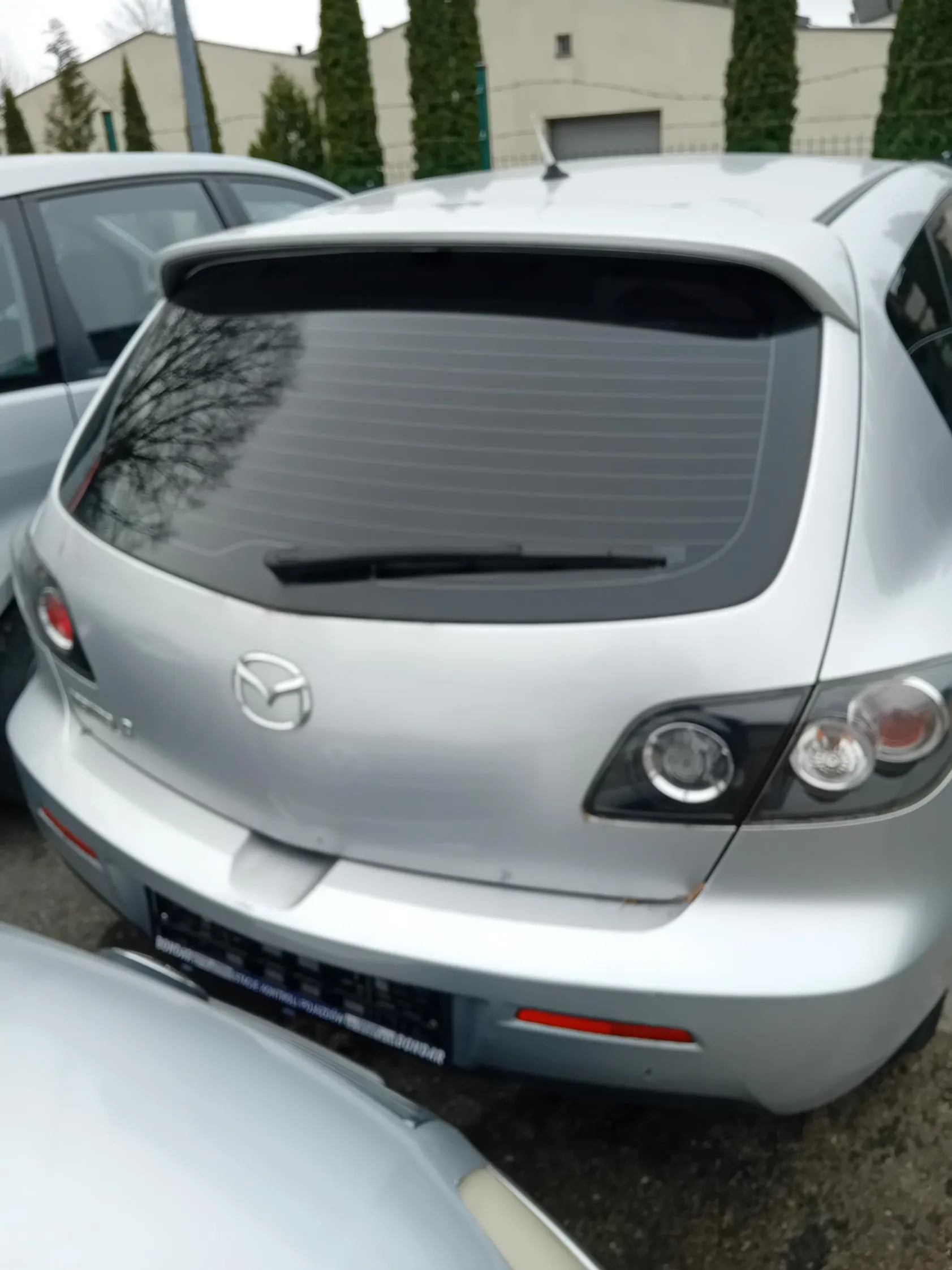 Tył srebrnego samochodu marki Mazda z widocznym logo, tylną szybą z wycieraczką, czerwonymi światłami tylnymi i tablicą rejestracyjną. W tle inne samochody, budynki oraz wysokie drzewa.