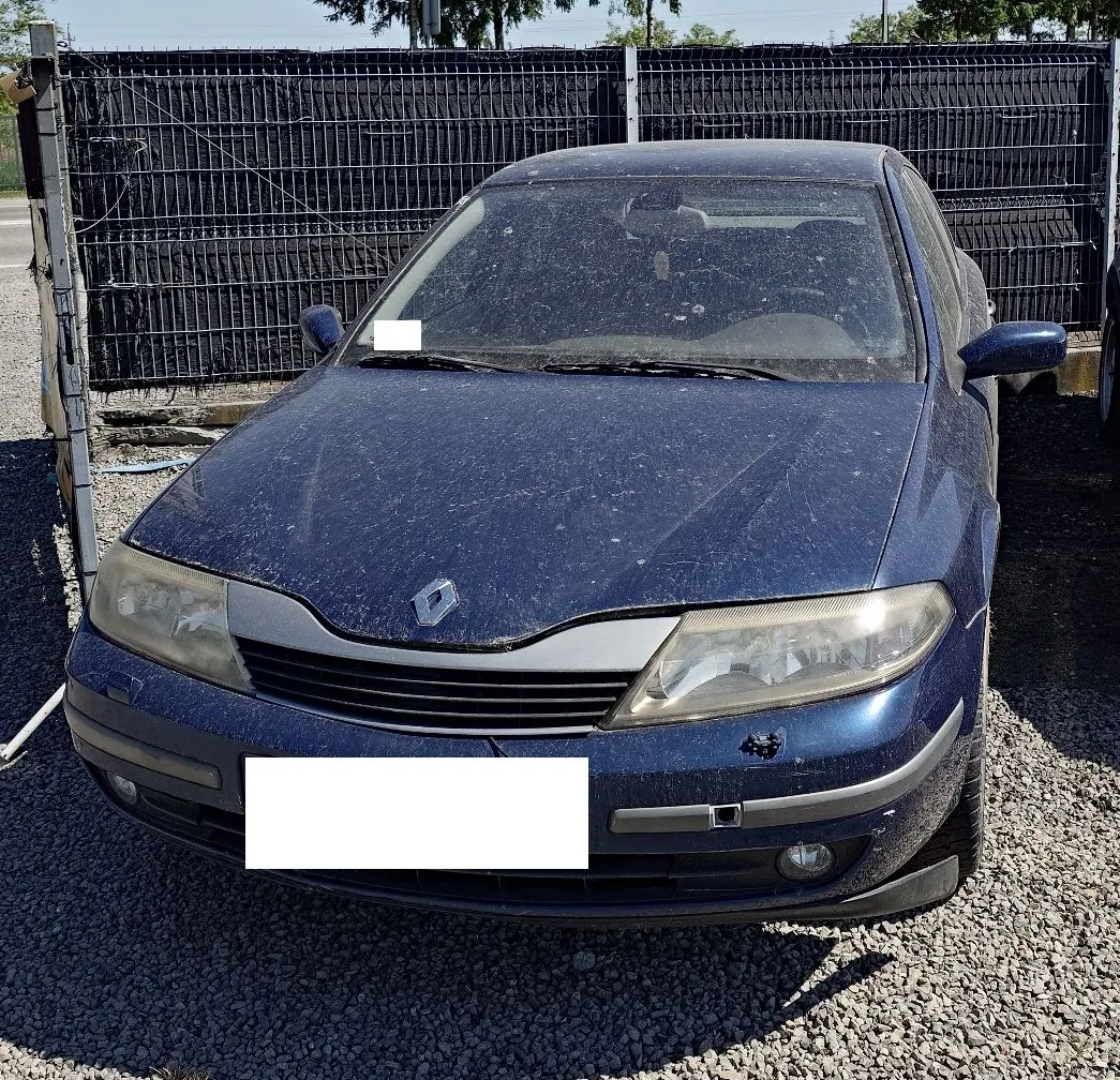 Zdjęcie przodu ciemnoniebieskiego samochodu marki Renault, zaparkowanego na żwirowym podłożu na tle czarnego ogrodzenia z siatki. Karoseria auta jest pokryta warstwą kurzu i drobnych zanieczyszczeń. Tablica rejestracyjna została zamaskowana białym prostokątem.
