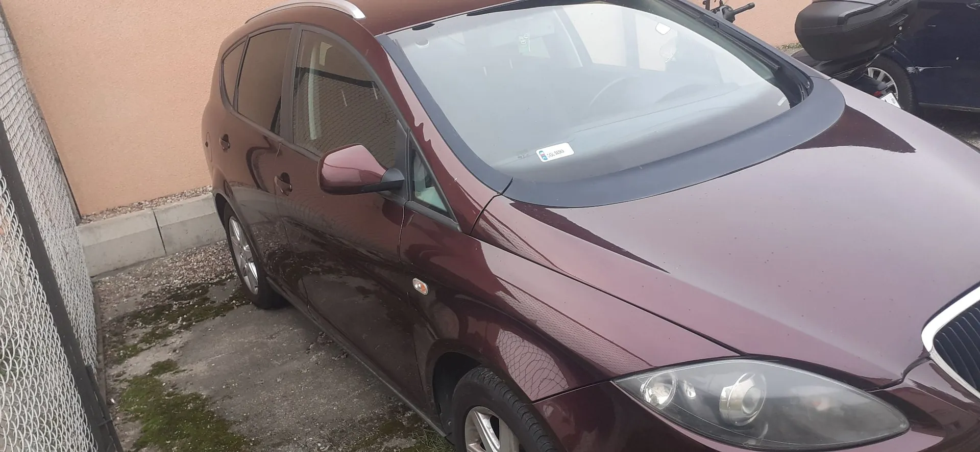 Bordowy samochód osobowy typu kombi zaparkowany obok jasnej ściany budynku i metalowego ogrodzenia z siatki. Widoczny jest przód i bok pojazdu, w tym reflektor, lusterko boczne oraz przednia szyba.
