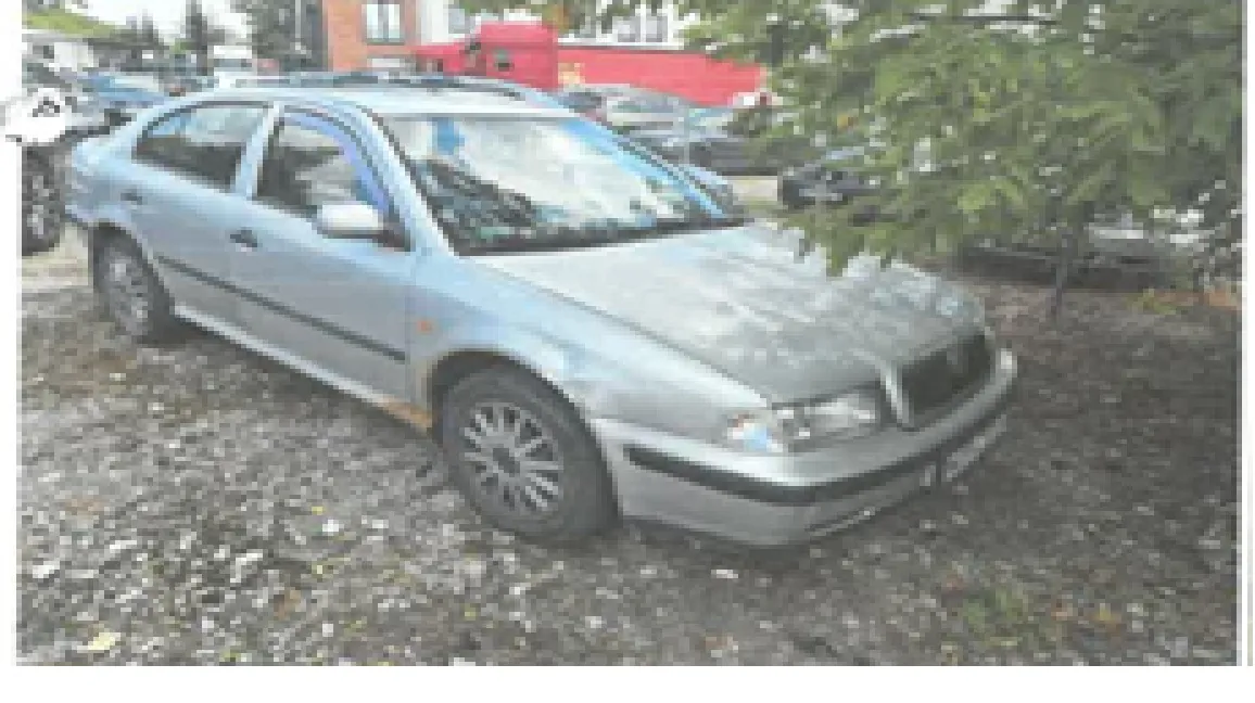 Srebrny samochód osobowy marki Skoda Octavia zaparkowany na żwirowym placu. Pojazd jest widoczny z przodu i z boku. W tle widać inne zaparkowane samochody, zielone drzewa oraz fragment czerwonego budynku.