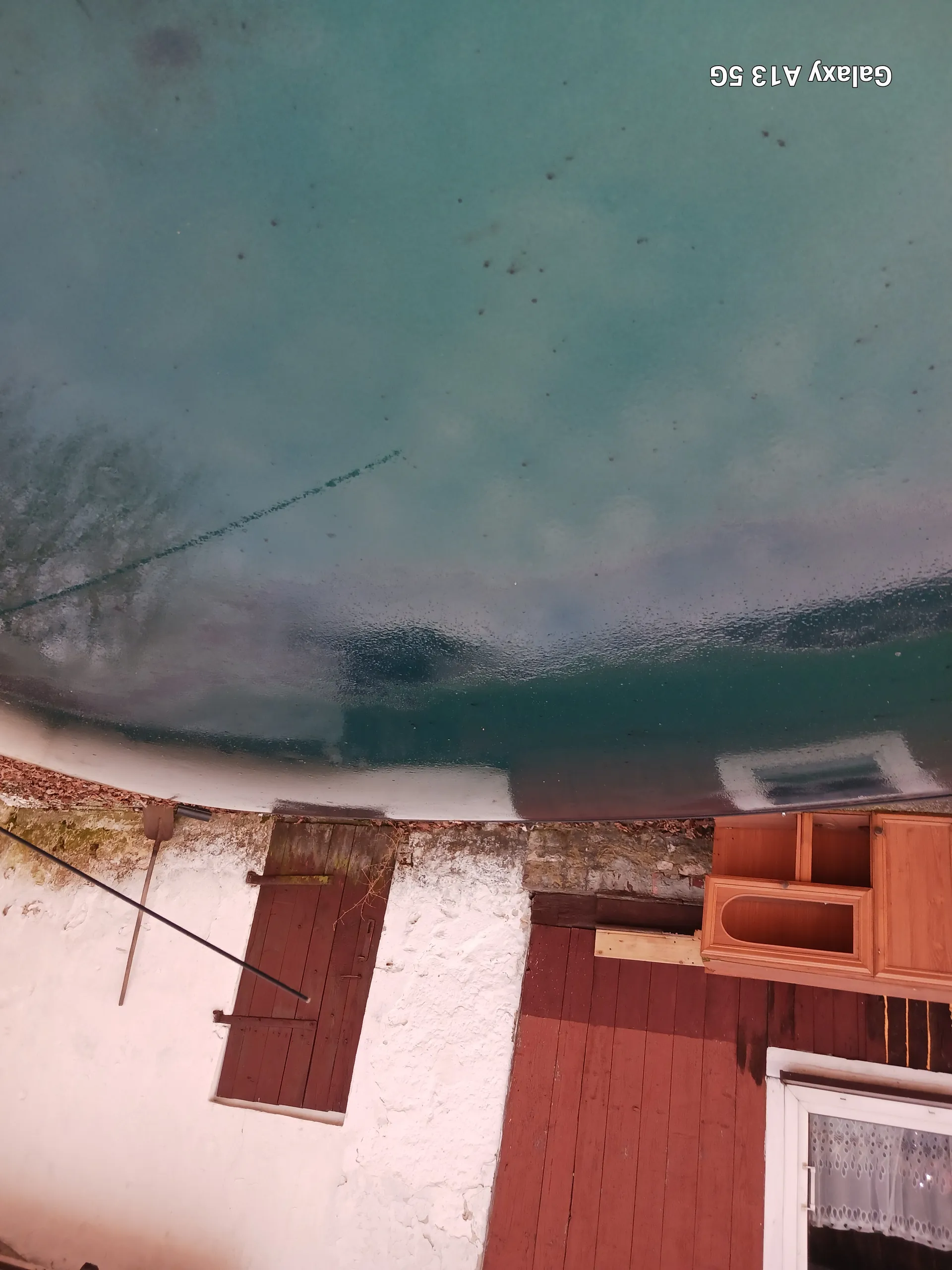 Duży cylindryczny zbiornik o niebiesko-zielonej powierzchni. Obok biały budynek z czerwonymi drewnianymi drzwiami i śniegiem na ziemi. Widoczna drewniana szafka oraz tekst 'Galaxy A13 5G' po prawej stronie obrazu.