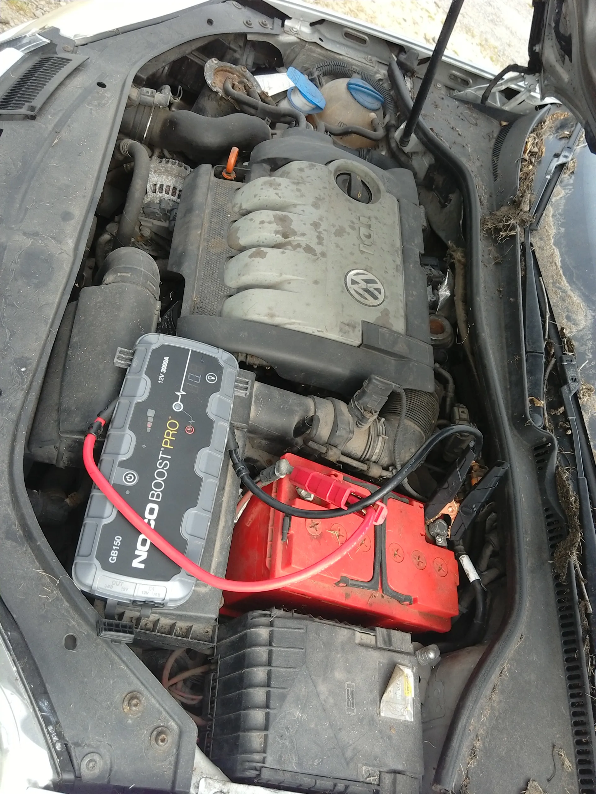 Widok otwartej tylnej części silnika samochodu. W centrum znajduje się czerwony akumulator podłączony czerwonym i czarnym kablem do urządzenia NOCO BOOST PRO GB150 oznaczonego jako 12V 3000A. Na pokrywie silnika widoczne są logo Volkswagena oraz napis TDI. Wokół znajdują się różne elementy silnika, w tym czarne rury, niebieskie nakrętki i zanieczyszczenia. Na lewo od akumulatora znajduje się czarna skrzynka z niewyraźnymi znakami.