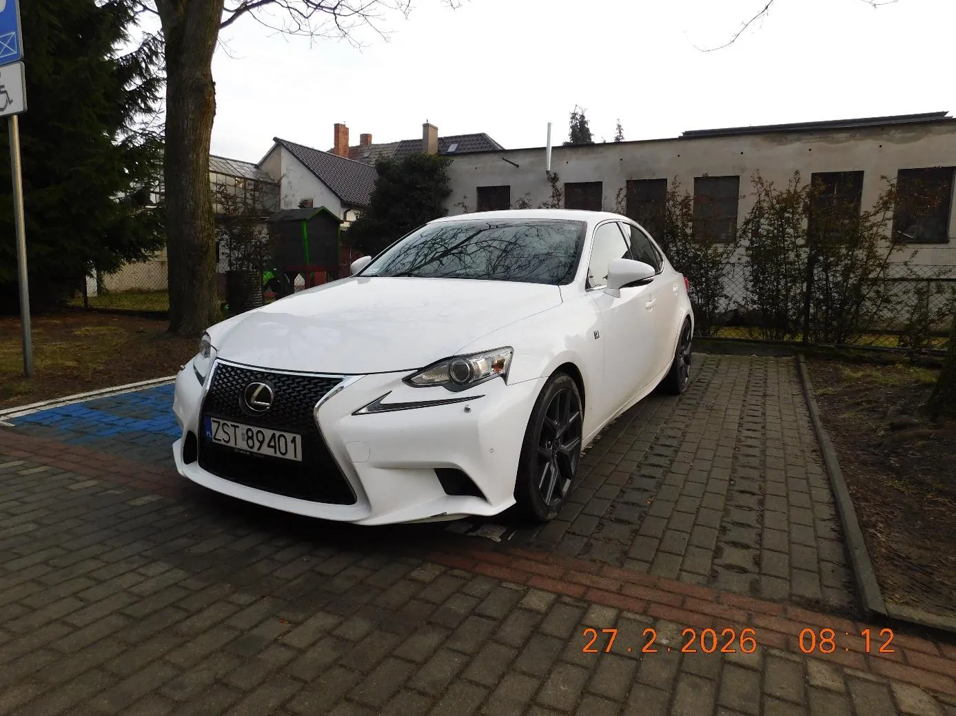 Biały samochód marki Lexus z tablicą rejestracyjną ZST 89401 zaparkowany na brukowanej powierzchni. W tle budynki, drzewa, krzewy oraz znak drogowy. W prawym dolnym rogu widoczny pomarańczowy znaczek z datą 27.2.2026 i godziną 08:12.
