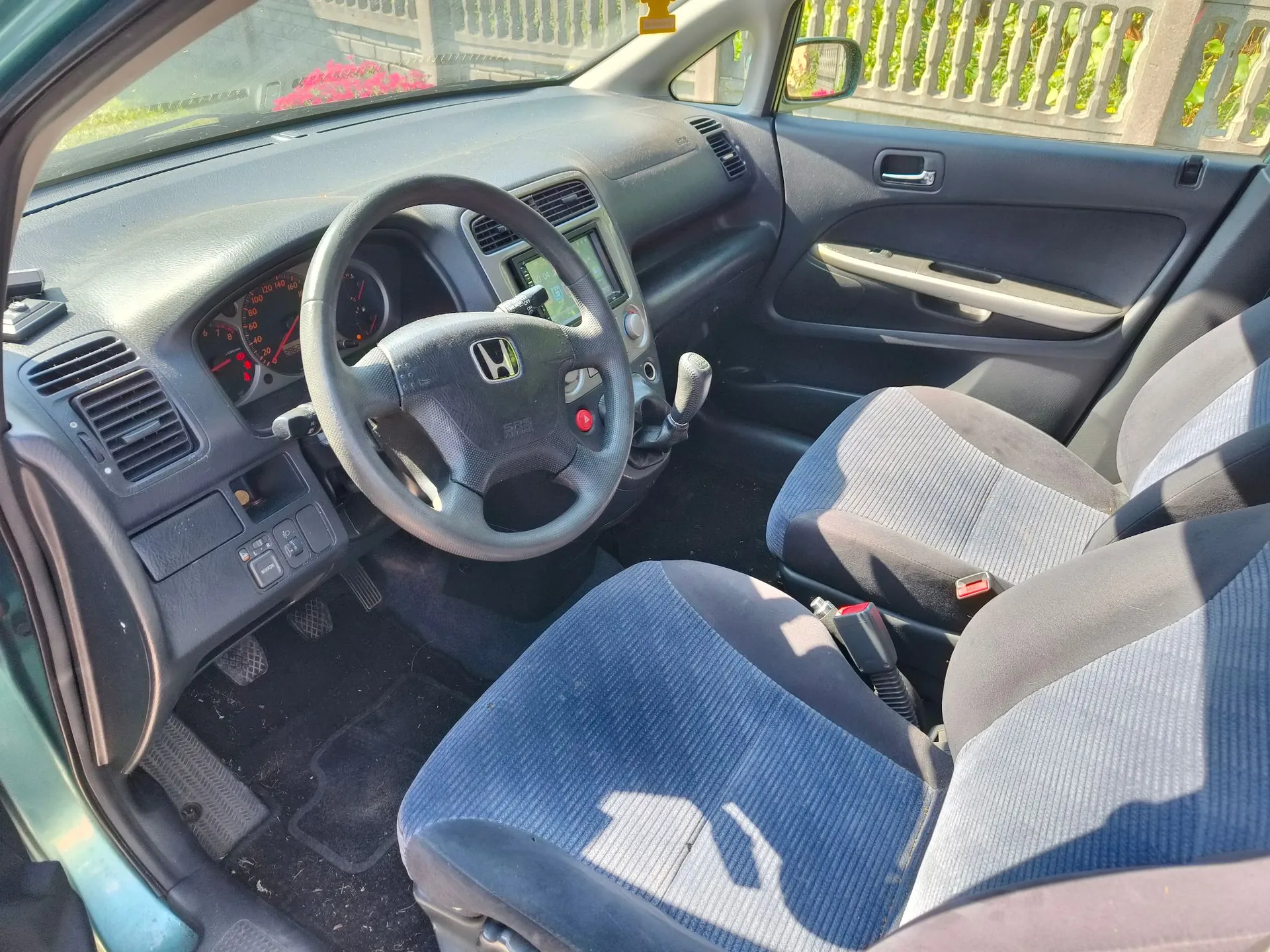Wnętrze samochodu marki Honda widziane z perspektywy pasażera. Widoczna jest kierownica z logo marki, deska rozdzielcza z zegarami, konsola środkowa z radiem oraz dwa przednie fotele obite niebieską tkaniną w prążki. Samochód ma kierownicę po lewej stronie.