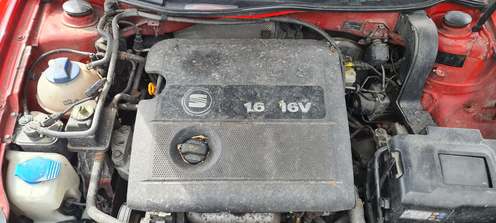 Widok z góry na zakurzoną komorę silnika w czerwonym samochodzie. W centrum znajduje się czarna plastikowa osłona silnika z logo marki Opel oraz napisem '1.6 16V'. Po lewej stronie widoczne są zbiorniki z płynami eksploatacyjnymi z niebieskimi zakrętkami, a po prawej czarna obudowa akumulatora.