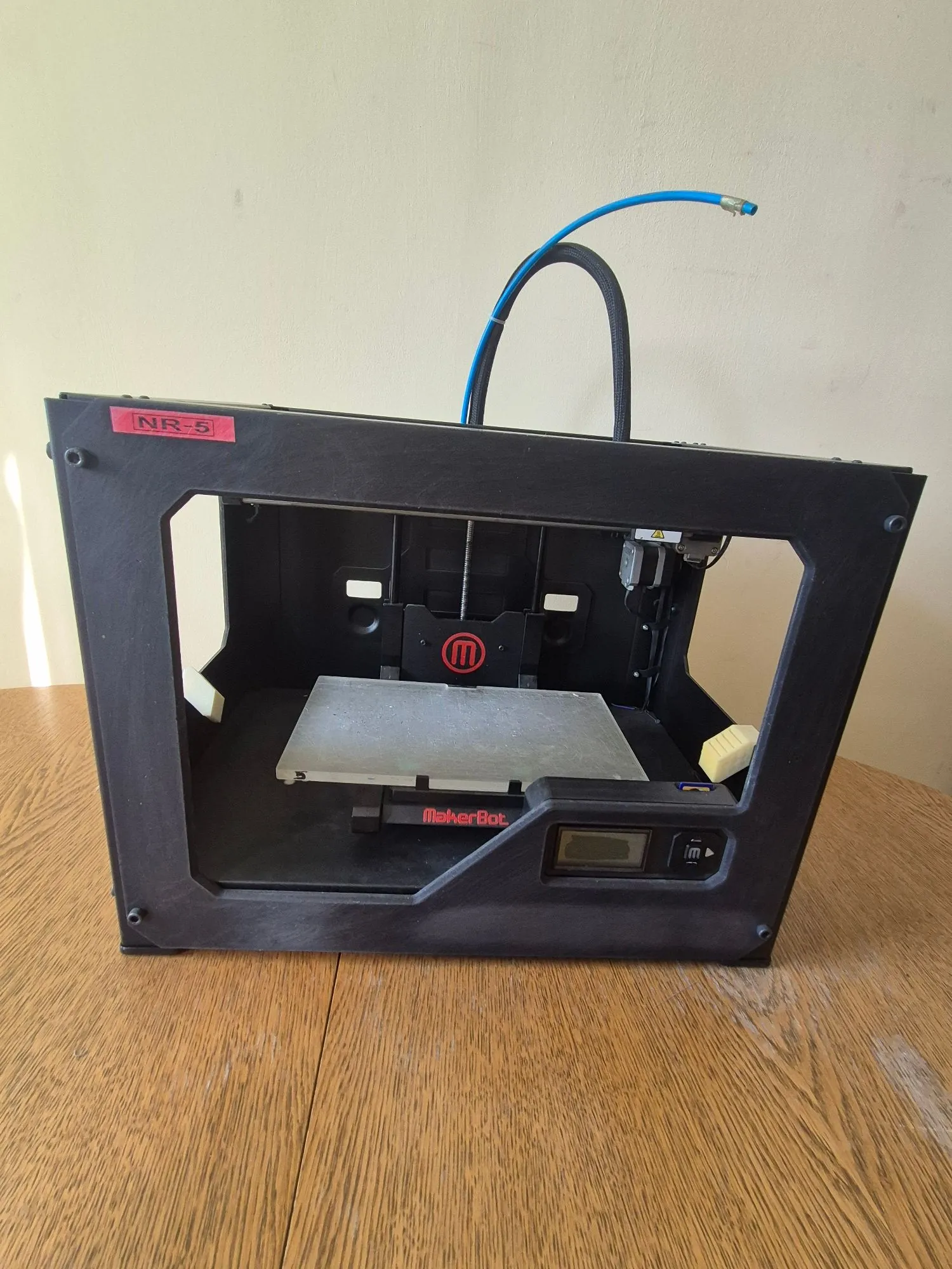 Czarna drukarka 3D marki MakerBot, model NR-5, ustawiona na drewnianym stole. Urządzenie posiada otwartą ramę, wewnątrz której znajduje się metalowa platforma robocza. Na przednim panelu widnieje napis 'MakerBot' oraz mały wyświetlacz LCD z przyciskami sterującymi. Z górnej części urządzenia wychodzi niebieski przewód.