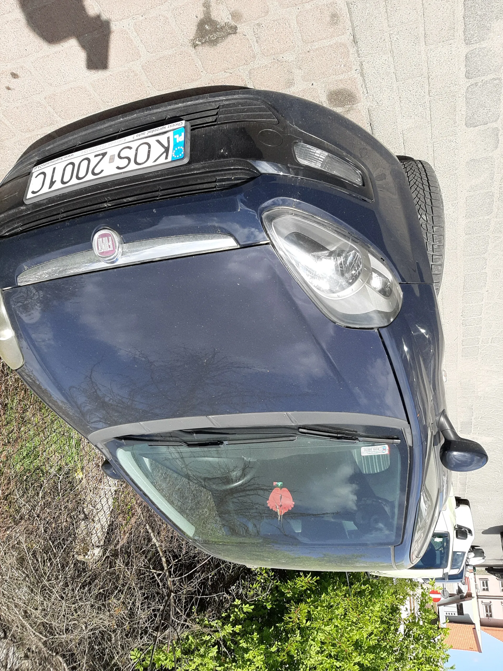 Ciemnoniebieski samochód marki Fiat z tablicą rejestracyjną KOS 2001C umieszczoną pionowo. Na przedniej szybie widoczna czerwona naklejka w kształcie serca. W tle zielone krzewy, suche gałęzie, budynki oraz brukowana powierzchnia. Samochód zaparkowany na otwartym obszarze.