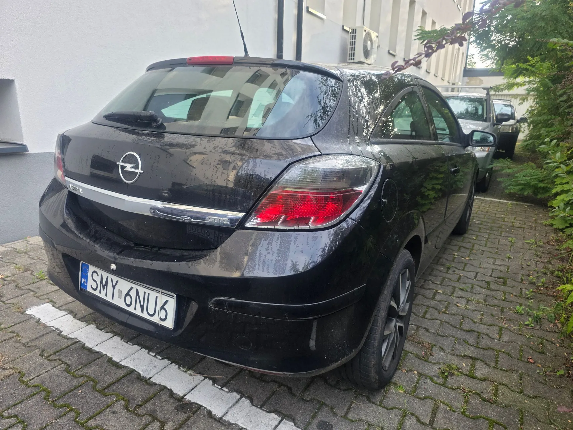 Tylny widok czarnego samochodu marki Opel zaparkowanego na kostce brukowej obok białego budynku. Samochód ma polską tablicę rejestracyjną o numerze SMY 6NU6. W tle widoczny jest inny, srebrny samochód oraz zielone krzewy.