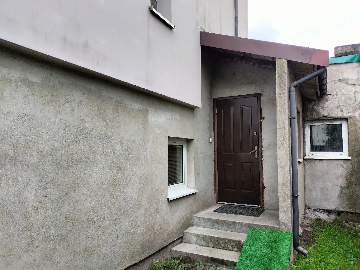 Widok na wejście do budynku z szarymi, tynkowanymi ścianami. Widoczne są ciemnobrązowe drzwi wejściowe z panelami, do których prowadzą trzy betonowe stopnie. Przed drzwiami leży ciemna wycieraczka, a obok schodów znajduje się zielona sztuczna trawa. Po lewej stronie drzwi jest małe, prostokątne okno, a po prawej rura spustowa odprowadzająca wodę z dachu. W tle widać fragment białej elewacji wyższego piętra.