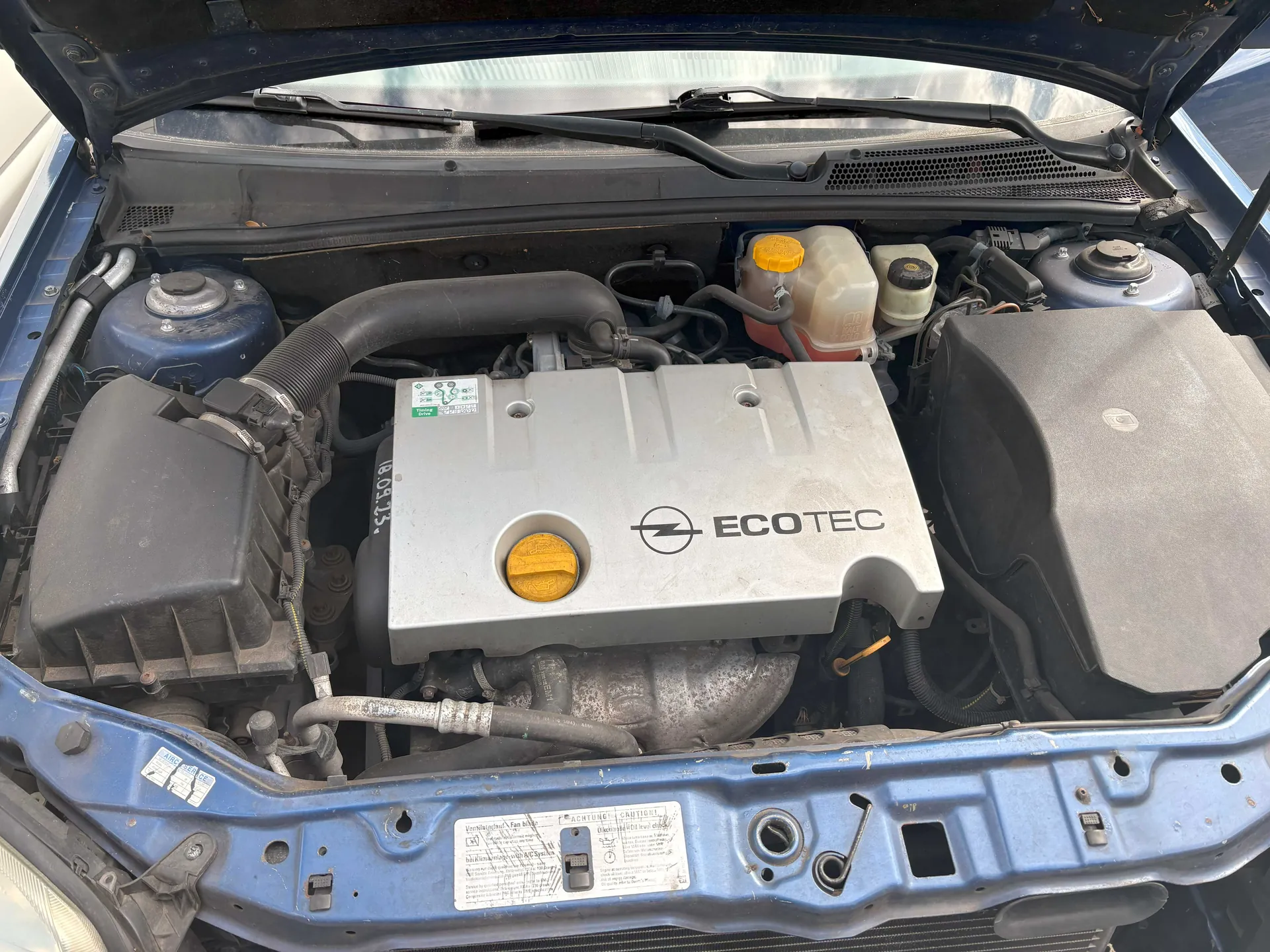 Widok otwartej tylnej części silnika samochodu z widocznym blokiem silnika oznaczonym napisem ECOTEC. W centrum znajduje się szara pokrywa silnika z żółtym korkiem, po prawej stronie zbiornik chłodniczy z żółtym korkiem i przezroczystą częścią z czerwoną cieczą. Po lewej stronie czarny filtr powietrza i niebieski zbiornik. Na dole widoczna niebieska ramka nadwozia z białą etykietą zawierającą tekst. Całość ukazana z góry, z otwartym kapotem.