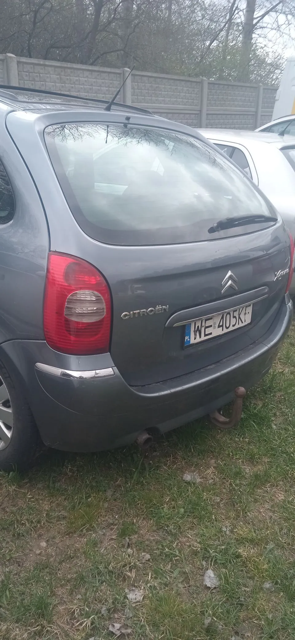 Tylny widok szarego samochodu marki Citroën z polską tablicą rejestracyjną o numerze WE 405KF. Pojazd posiada zamontowany hak holowniczy i jest zaparkowany na trawiastym poboczu obok betonowego ogrodzenia.