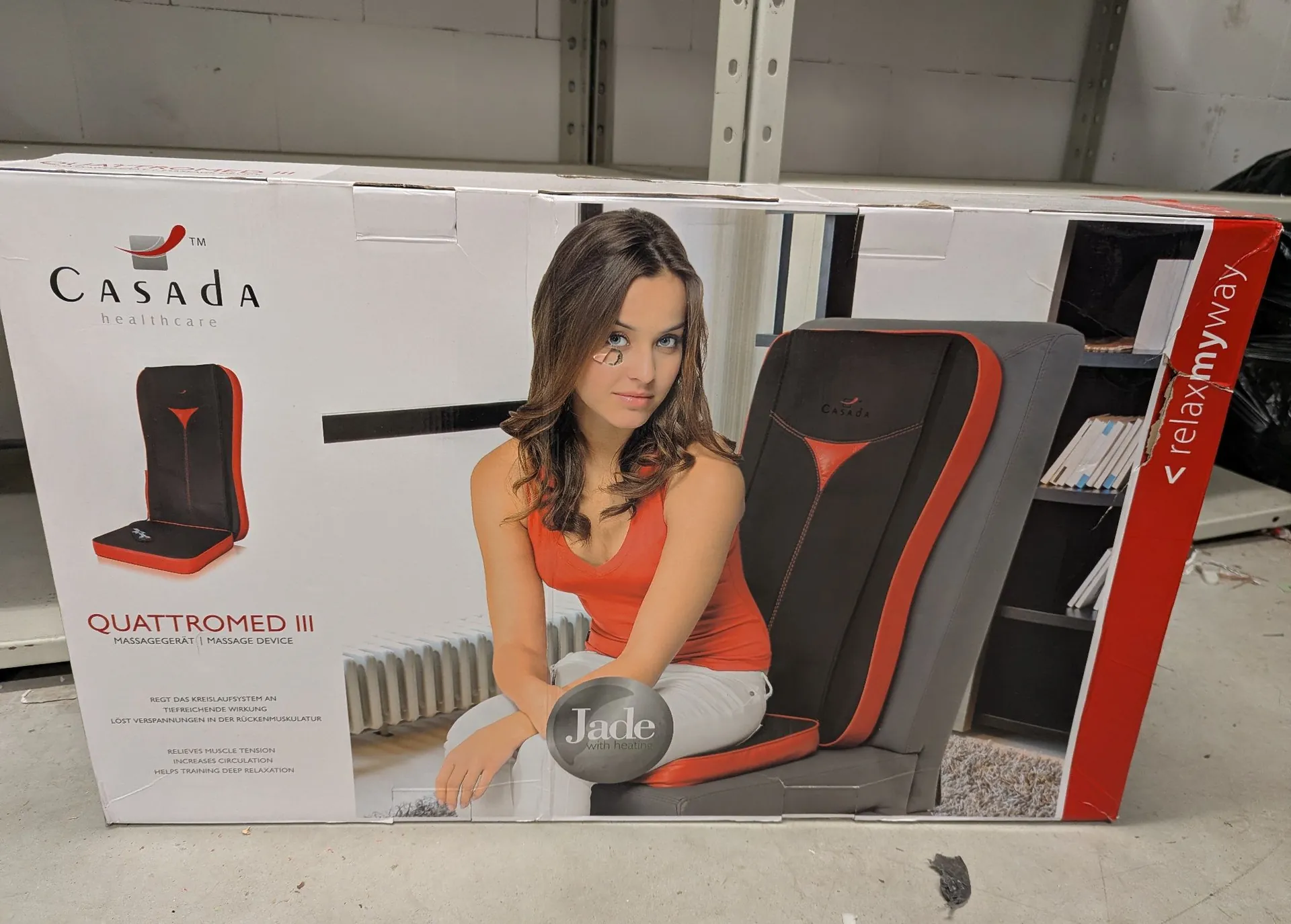 Kartonowe opakowanie urządzenia do masażu marki Casada Healthcare. Na pudełku widnieje napis 'CASADA healthcare' oraz 'QUATTROMMED III MASSAGEGERÄT | MASSAGE DEVICE'. Przedstawiona jest kobieta w pomarańczowej koszulce i białych spodniach siedząca na urządzeniu do masażu. Po lewej stronie znajduje się mniejszy obrazek produktu, a w dolnej części okrągła naklejka z napisem 'Jade with Heat'. Po prawej stronie widnieje napis 'relaxmyway'.