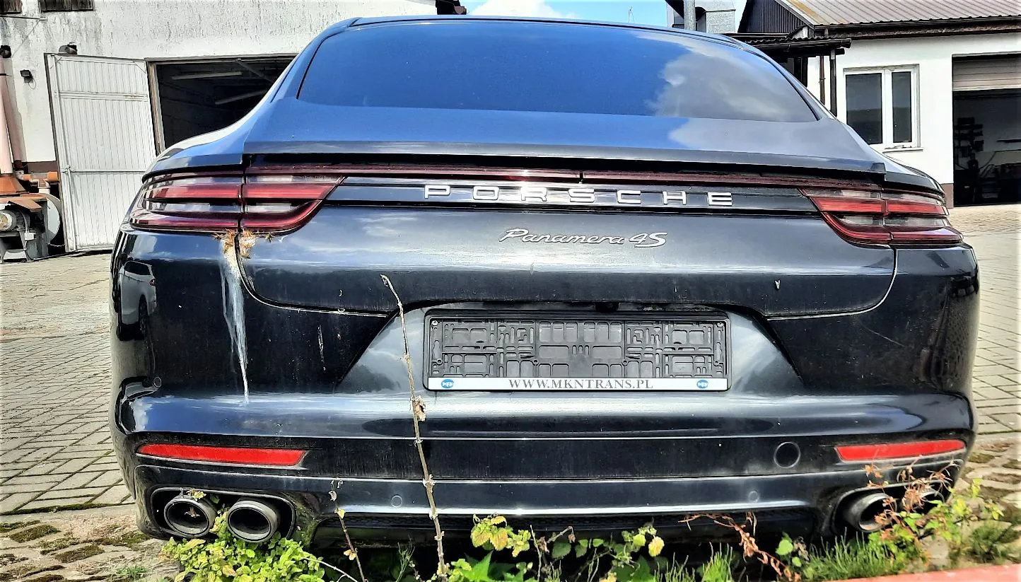 Tylny widok ciemnoniebieskiego samochodu marki Porsche Panamera 4S. Na klapie bagażnika widnieje napis 'PORSCHE' oraz model 'Panamera 4S'. Samochód nie posiada tablic rejestracyjnych, a w ramce widnieje adres strony internetowej 'WWW.MKNTRANS.PL'. Pojazd jest zabrudzony, widoczne są zacieki i pył na karoserii. Na dole znajdują się podwójne rury wydechowe po obu stronach. Samochód stoi na kostce brukowej, a u dołu widoczne są zielone rośliny.
