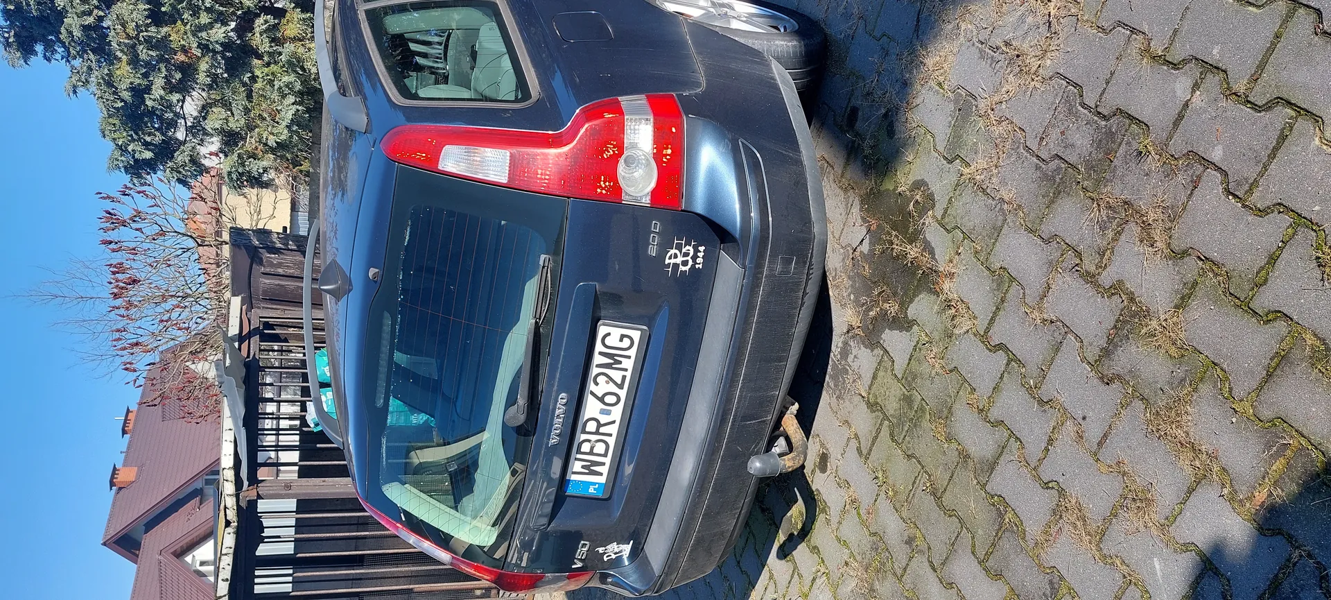 Z tyłu ciemnoniebieski samochód Volvo V50 z tablicą rejestracyjną WBR 62MG (PL), widocznym holownikiem, naklejkami z napisami 200 i 1944 oraz napisem VOLVO. Samochód zaparkowany na brukowanej powierzchni z trawą między kamieniami. W tle domy z czerwonymi dachami, ogrodzenie i drzewa pod jasnym niebem.
